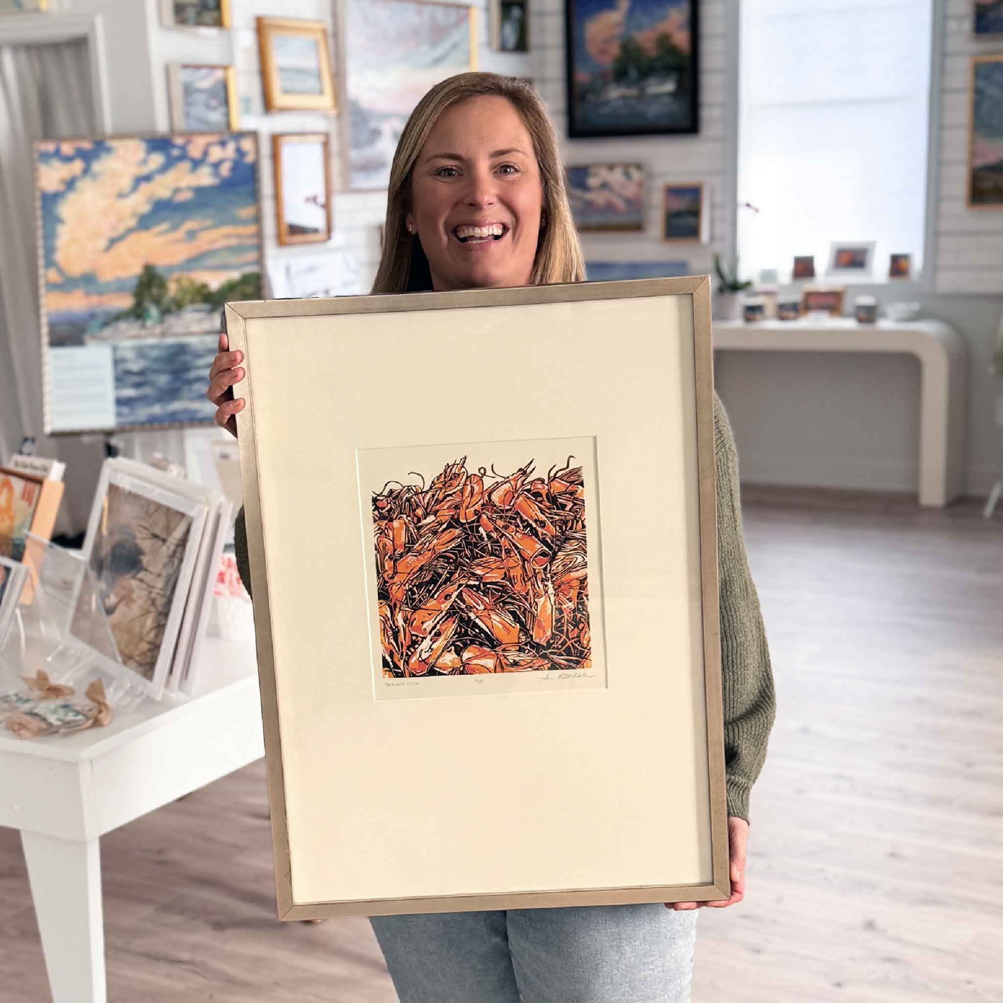 Bordy at Gray Man Gallery holding framed “Shrimp Stock” original linocut  by Laurie McIntosh featuring bold orange shrimp pile artwork, coastal Lowcountry seafood scene.