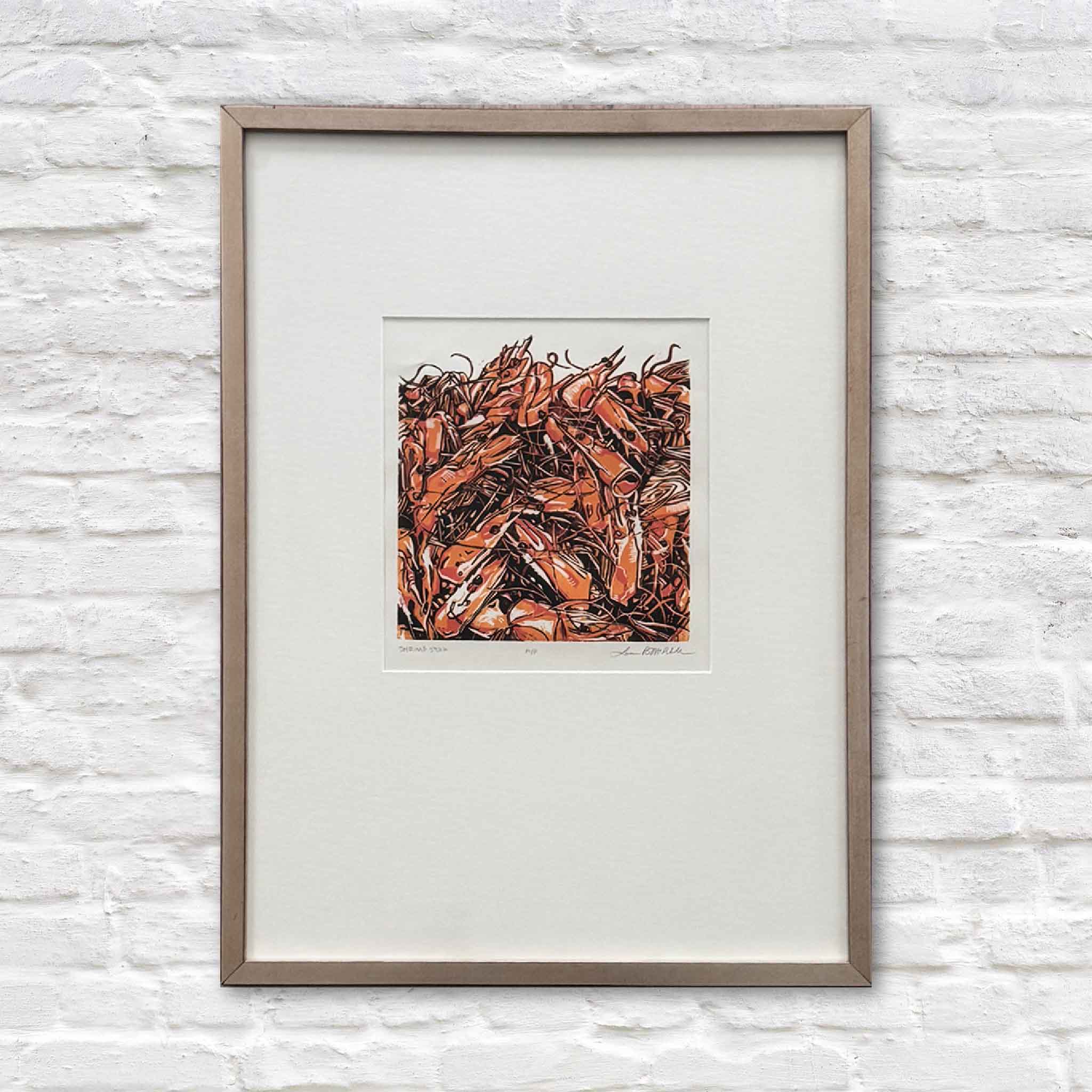 Framed “Shrimp Stock” original linocut art by Laurie McIntosh featuring bold orange shrimp pile artwork, coastal Lowcountry seafood scene, hanging on white brick wall at Gray Man Gallery Pawleys Island.