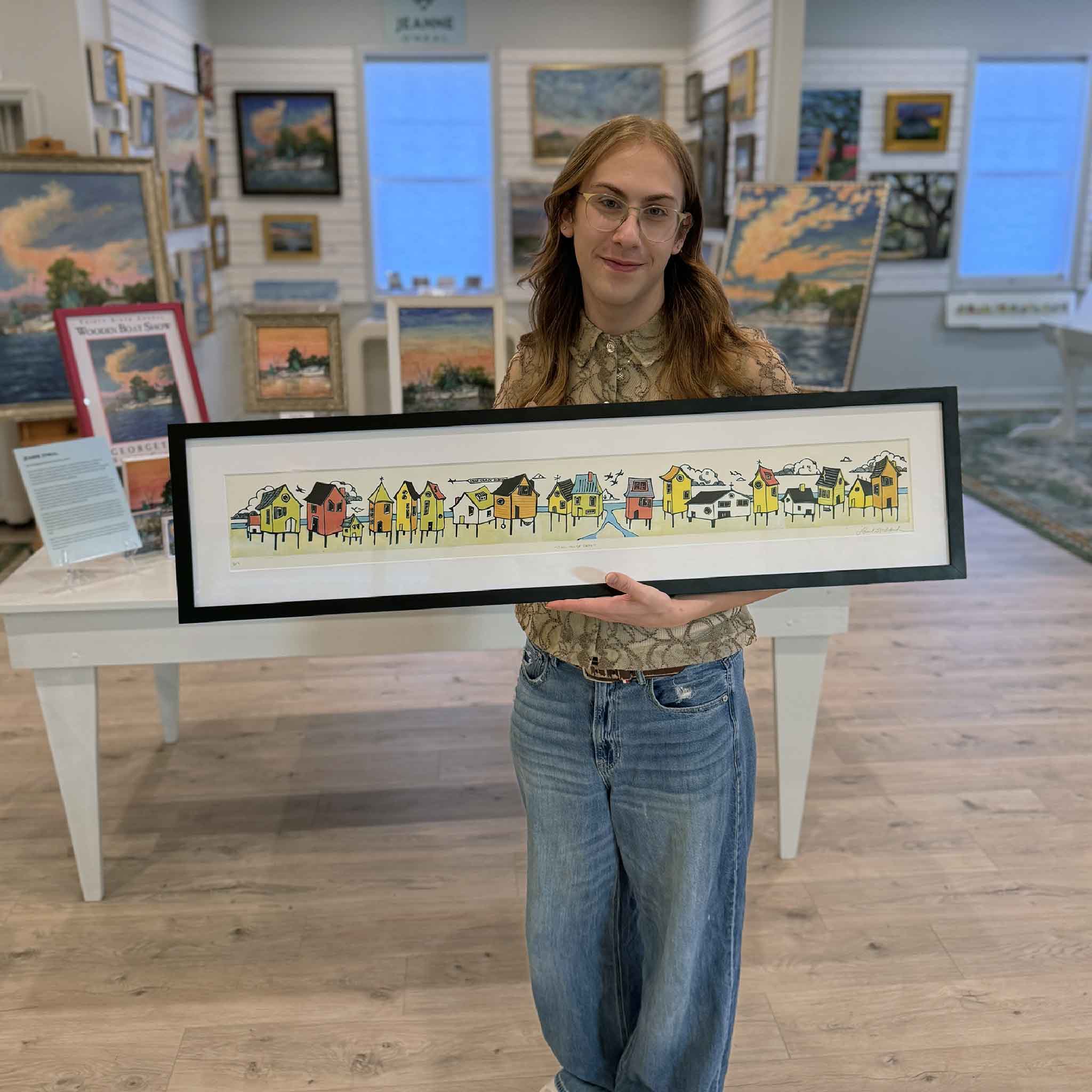 Gallery team member Alex displaying Stick House Beach by Laurie McIntosh at Gray Man Gallery. This original linocut reduction print showcases a cheerful collection of colorful beach houses, coastal wildlife, and playful architectural details inspired by the South Carolina coast and Lowcountry beach communities.