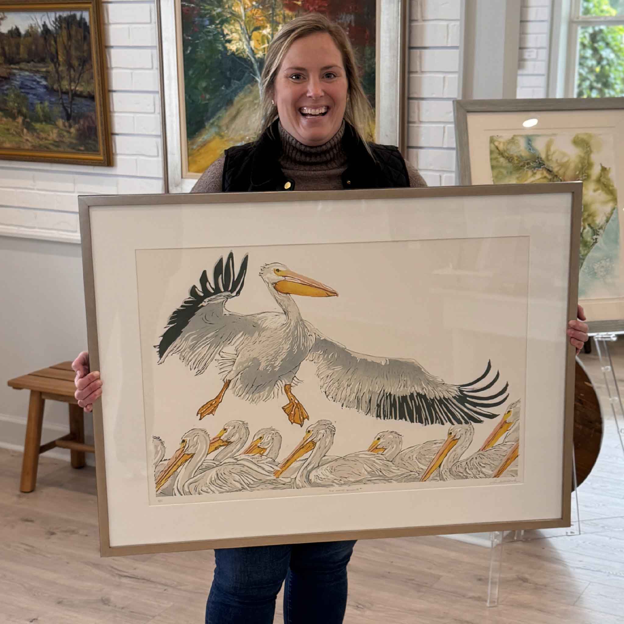 Local South Carolina artist Laurie McIntosh holding her original linocut print “The White Pelicans” at Gray Man Gallery in Pawleys Island, featuring white pelicans in flight, framed coastal wildlife artwork inspired by the Lowcountry

