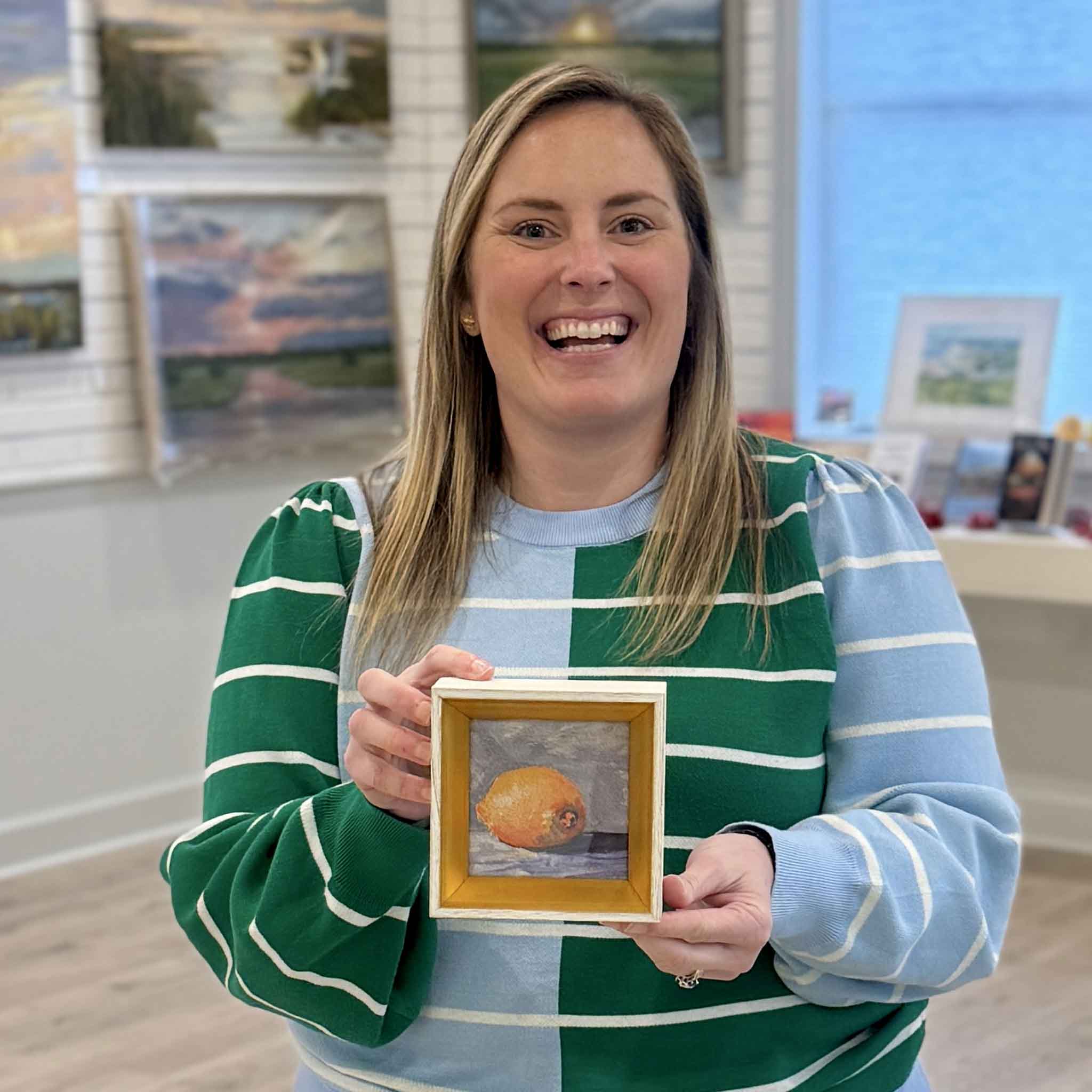 Bordy holds “Lemon Drop” by Jeanne O’Neal inside Gray Man Gallery in Pawleys Island, South Carolina. The small framed lemon art print features a classic gold frame and painterly citrus still life, showcasing the scale and depth of this Lowcountry local artist piece available at Gray Man Gallery.