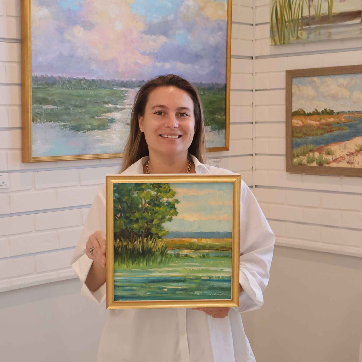 A Gray Man Gallery team member holding Coastal Tides, a framed original oil painting by local artist Libby Smart, inside the art gallery in Pawleys Island, South Carolina. The intimate Lowcountry marsh landscape features lush green trees, blue and turquoise tidal waters, golden marsh grasses, and a soft coastal sky, with the photograph showing the painting's scale and elegant gold frame.