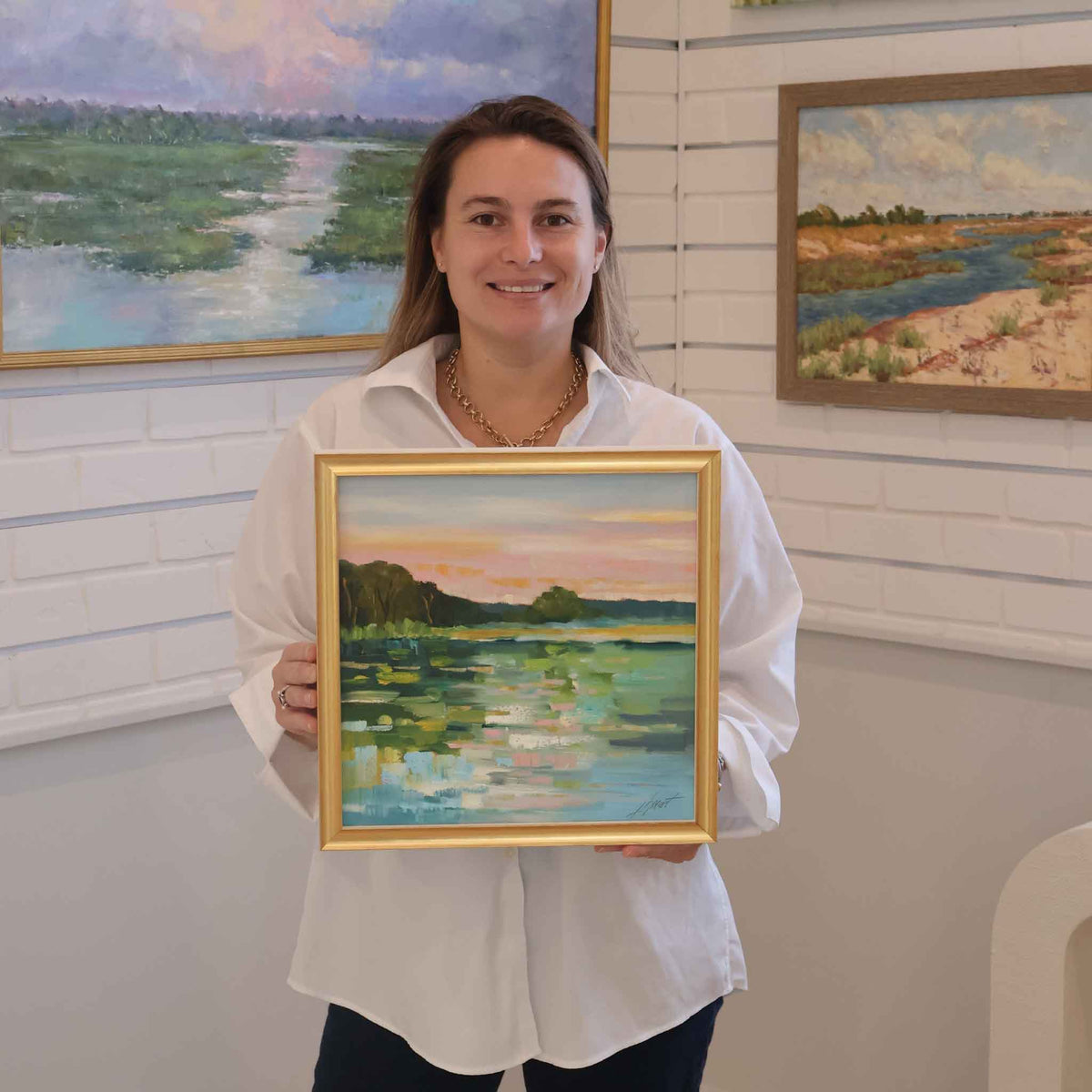 Gray Man Gallery team member holding Glistening Water, a framed original oil landscape painting by local artist Libby Smart, inside the gallery in Pawleys Island, South Carolina. The colorful coastal waterscape features sparkling blue-green water, dark shoreline trees, soft pink and peach clouds, and an elegant gold frame, with the photograph showing the artwork's scale and presentation.