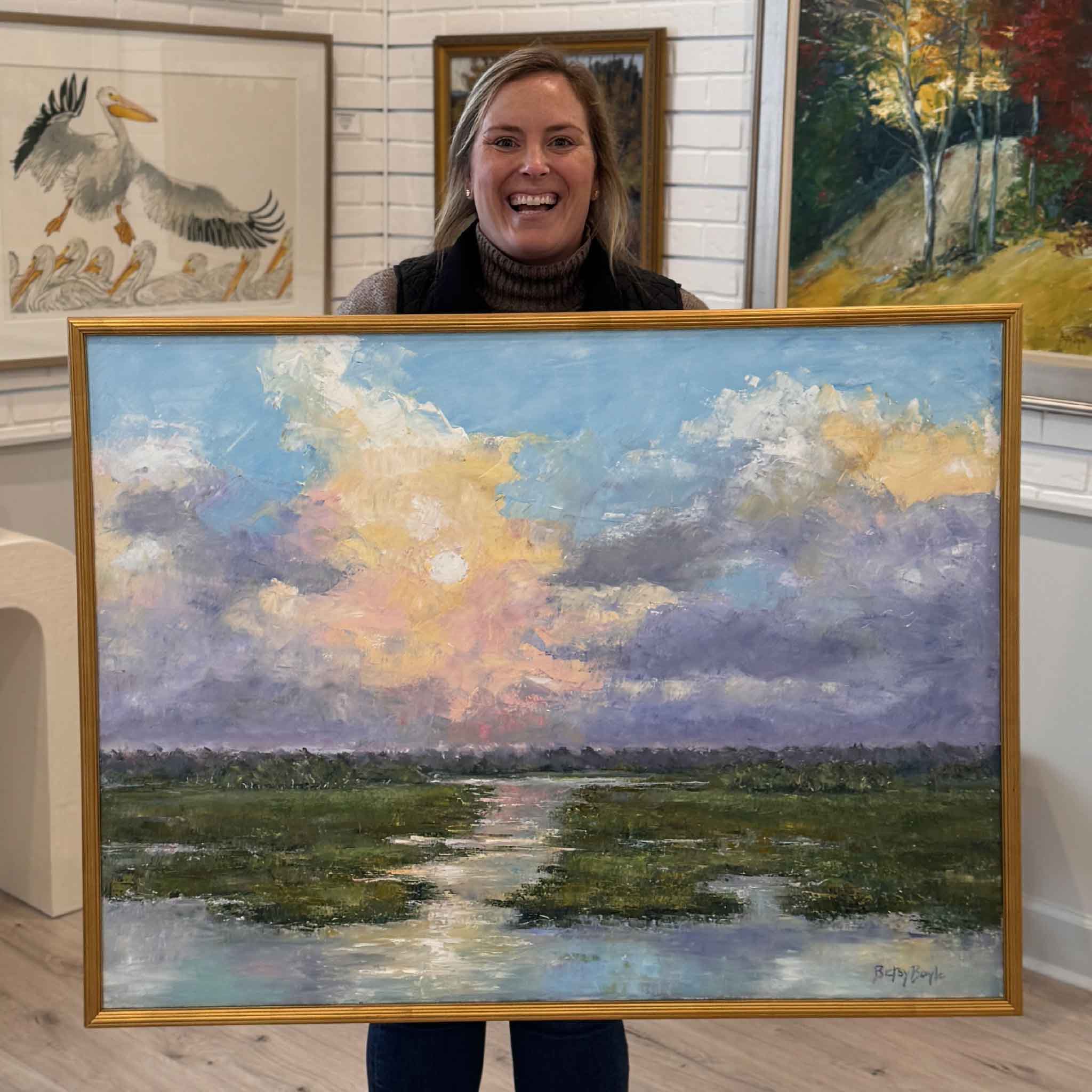 Bordy holding the framed original oil painting “Light Overcomes” by local Lowcountry artist Betsy Boyle inside Gray Man Gallery in Pawleys Island, South Carolina, showcasing a luminous coastal marsh landscape with pastel clouds, reflective water, and expressive brushwork, custom framed in gold and professionally finished by Gray Man Gallery, highlighting fine art craftsmanship, local artists, and boutique coastal gallery presentation