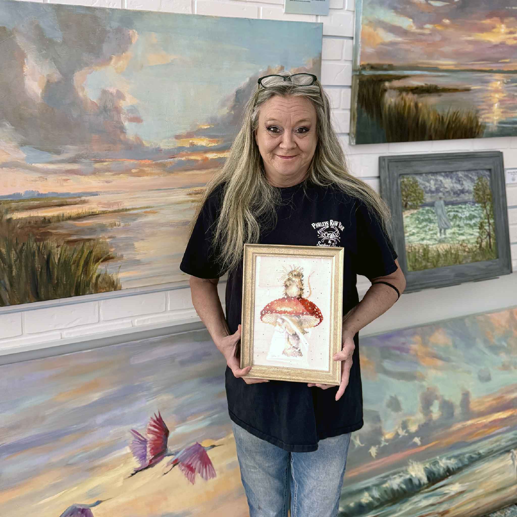 A Gray Man Gallery team member holds Little Forager by Mimi Beaver, a framed cross-stitch artwork donated to benefit Salty Dogs Senior Animal Rescue, with all proceeds supporting senior dogs.