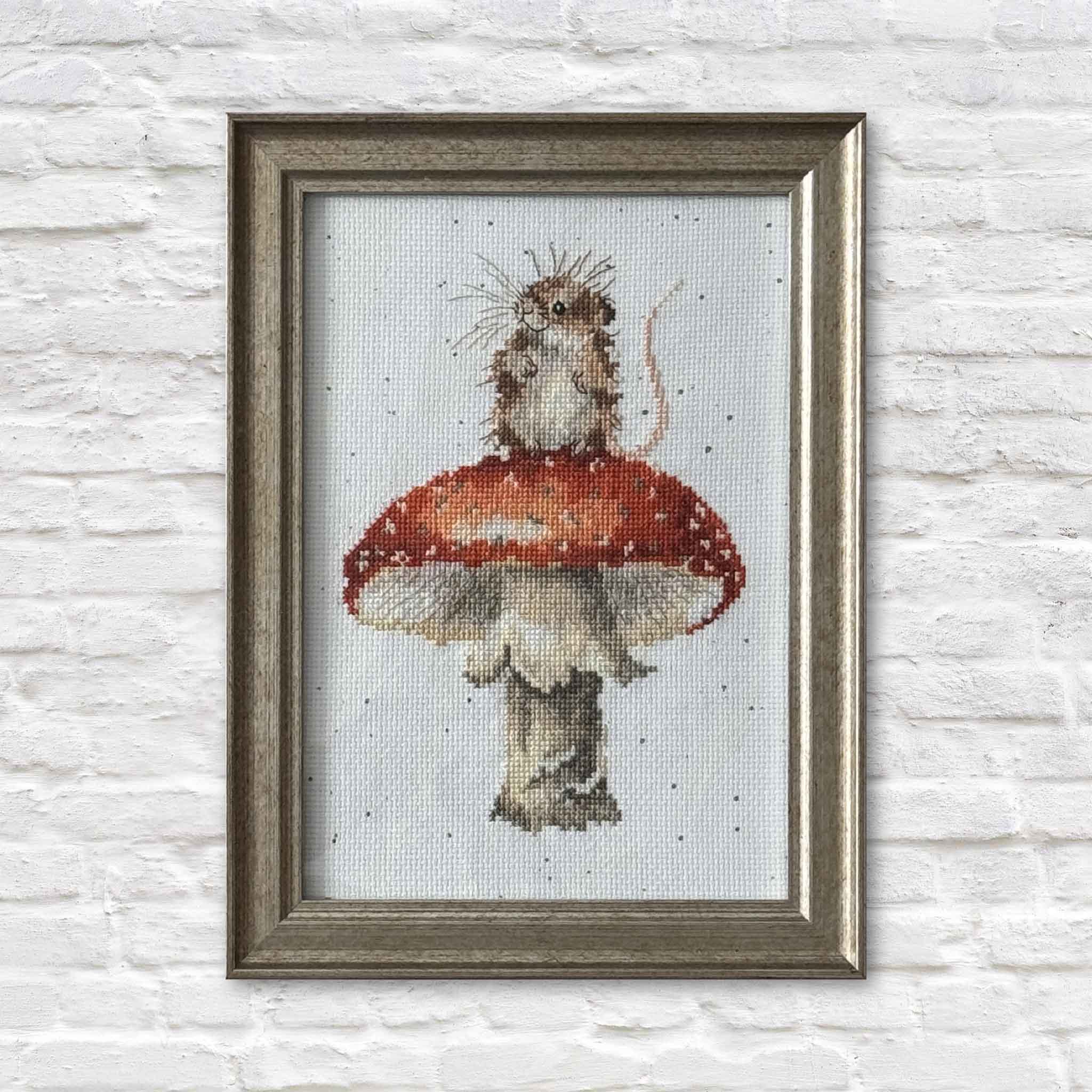 An original framed cross-stitch by Mimi Beaver showing a small mouse standing on a red mushroom, stitched in soft neutral tones on a light fabric background.