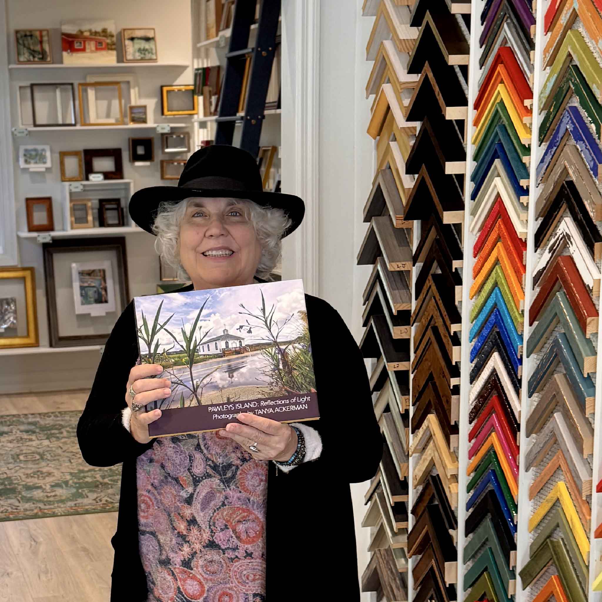 Local Pawleys Island photographer Tanya Ackerman holding her Reflections of Light photography book inside Gray Man Gallery surrounded by custom frame samples