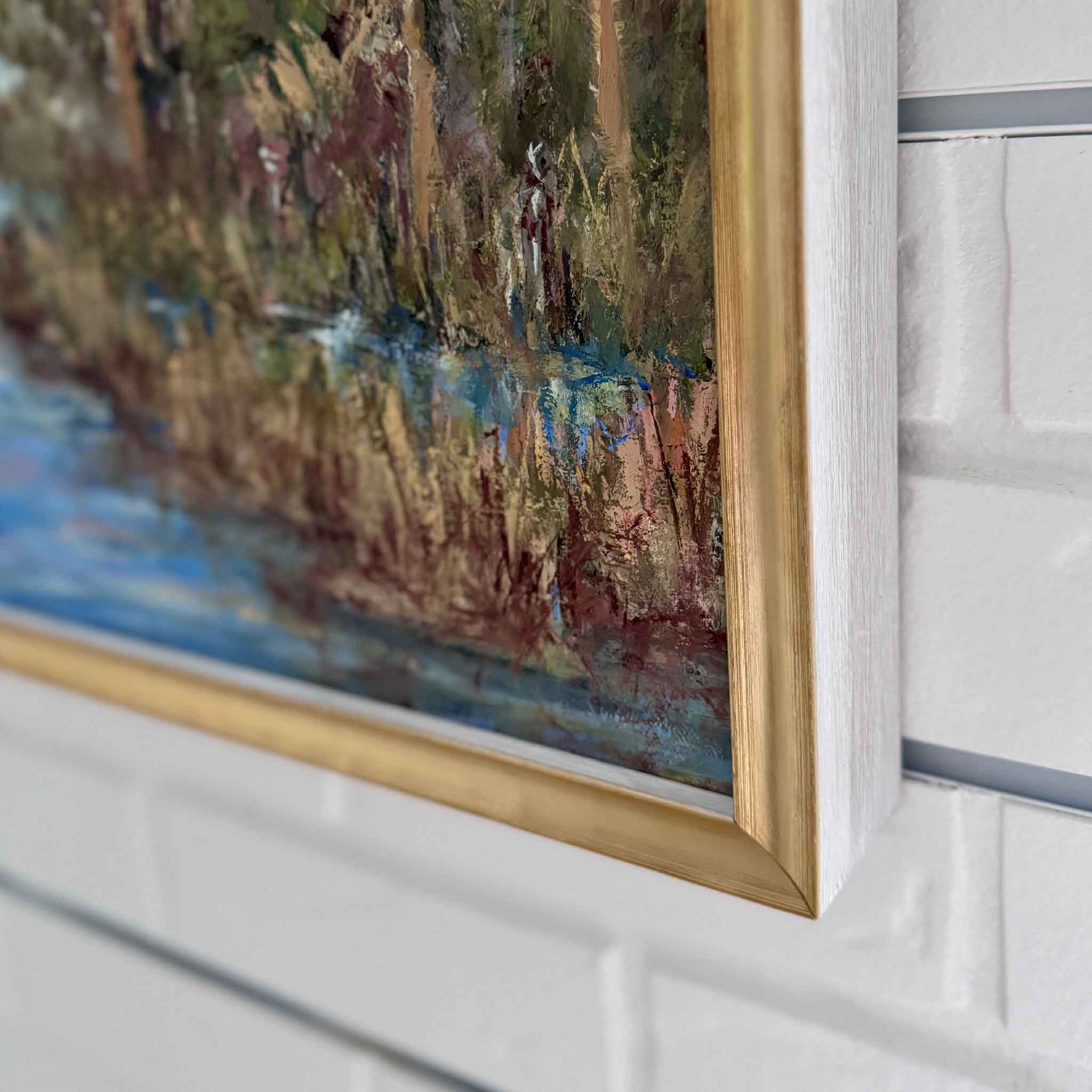 Framed painting of a natural landscape with trees and water on a white wall.