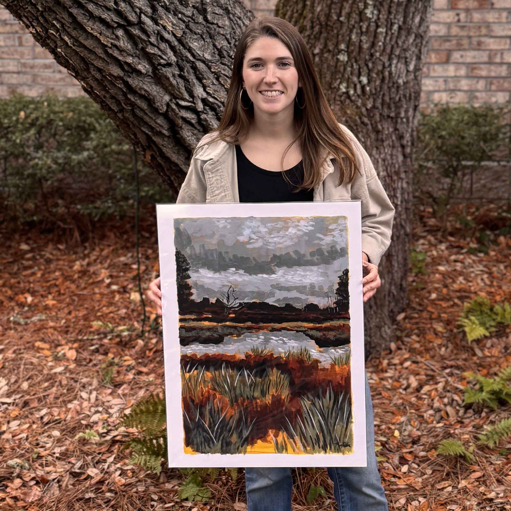 Maggie Pelton, local artist, holding her Oyster Landing Basin fine art print outdoors, depicting layered marsh grasses and reflective tidal waters inspired by North Inlet and the Georgetown Lowcountry, available at Gray Man Gallery in Pawleys Island, South Carolina