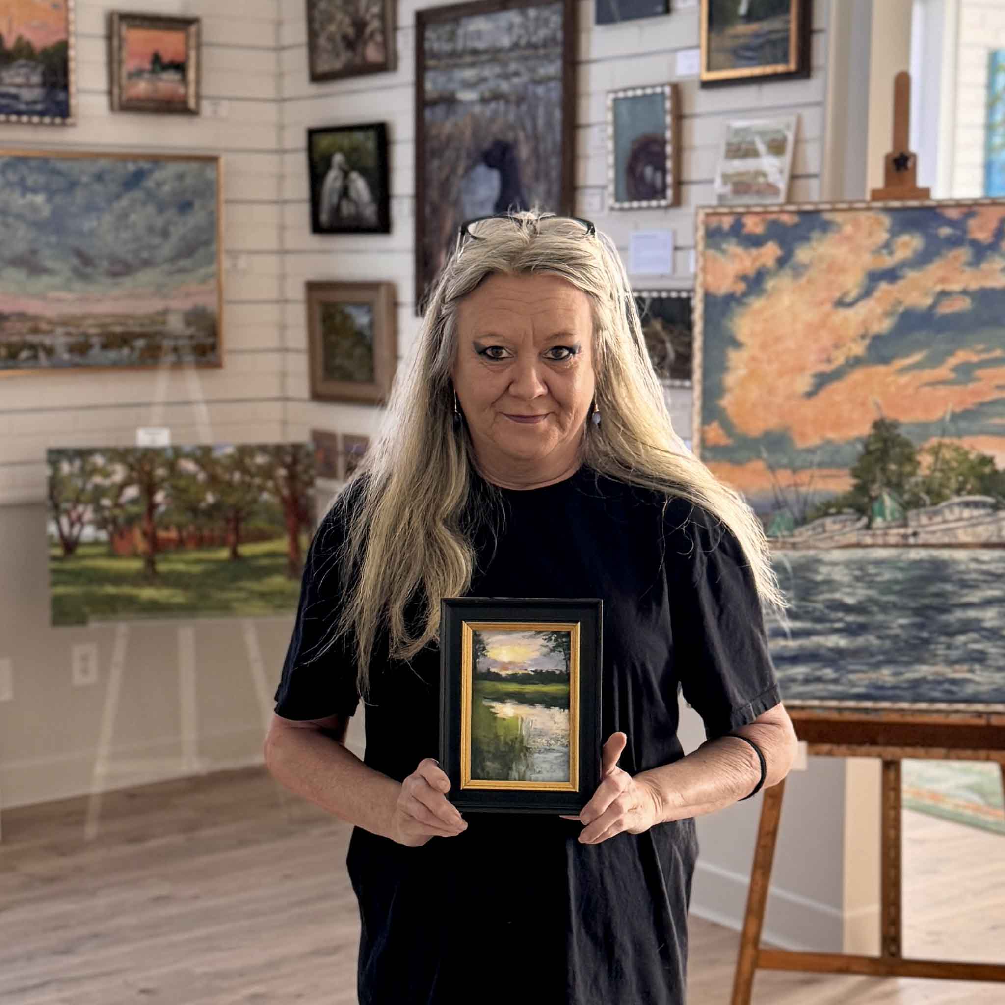 Artist Joanne Willoughby holds Marsh Sunset, a 4x6 original pastel framed and ready to display, capturing the quiet beauty of a Lowcountry marsh at sunset inside Gray Man Gallery.