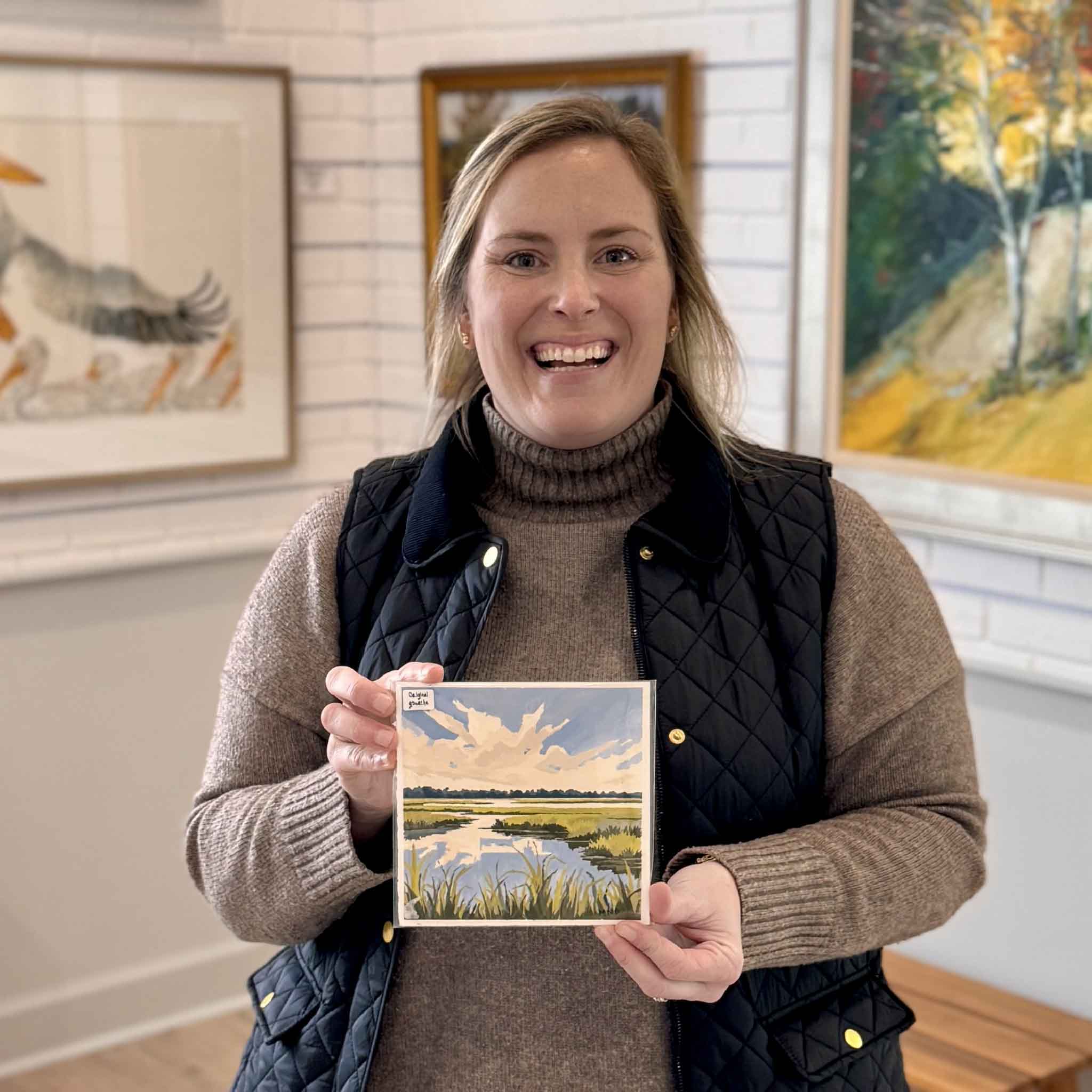 A Gray Man Gallery team member holds “Marsh View I,” an original 6x6 gouache painting by local Lowcountry artist Megan DuMont, featuring a coastal marsh landscape with winding tidal creeks, golden grasses, and dramatic clouds, photographed inside Gray Man Gallery in Pawleys Island, South Carolina, showcasing handcrafted fine art and custom framing