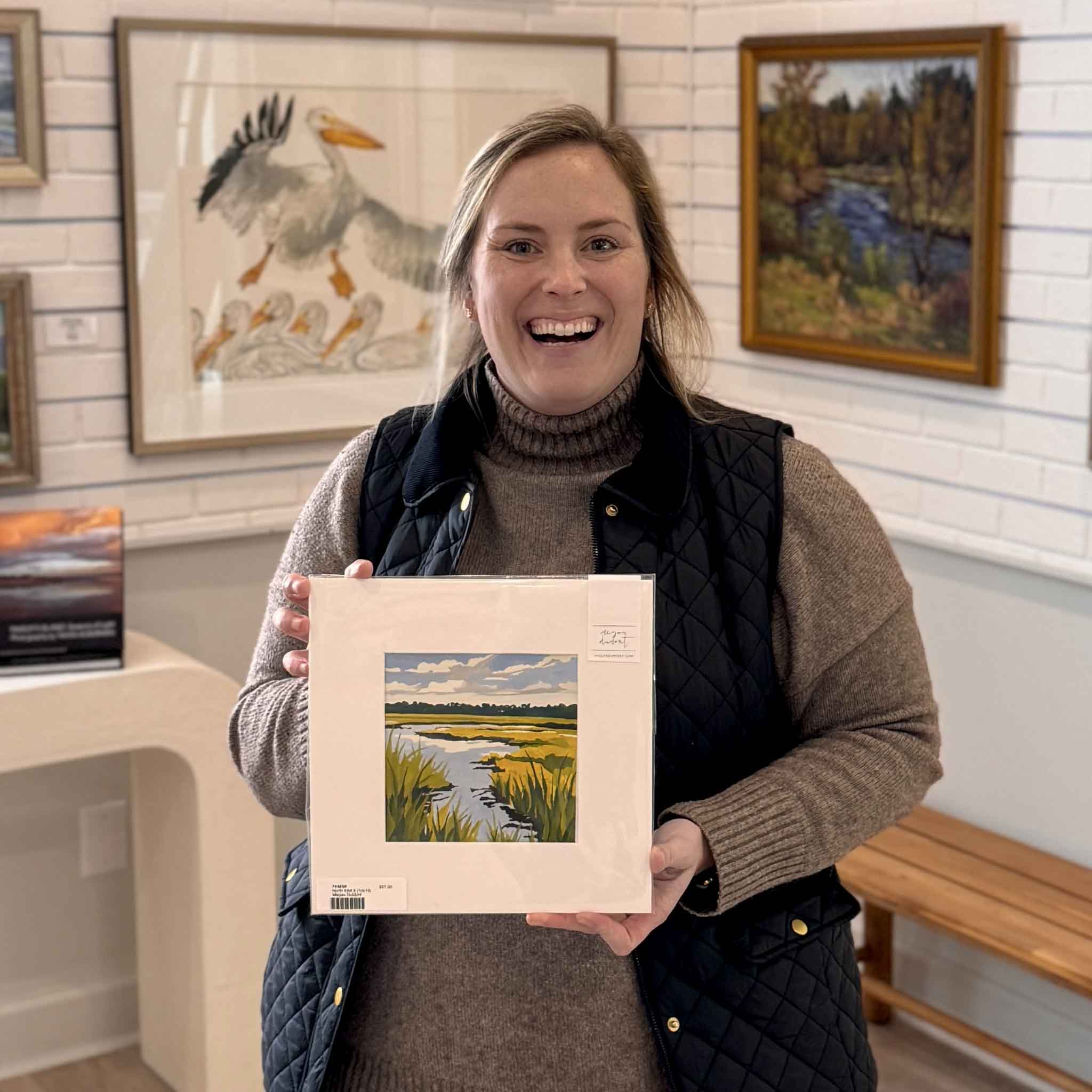 A smiling Gray Man Gallery team member holds Megan DuMont’s “North Inlet II,” an original 10x10 gouache painting of the South Carolina Lowcountry marsh, featuring winding tidal creeks, golden grasses, soft blue water, and cloud-filled skies, displayed inside Gray Man Gallery in Pawleys Island with custom-framed coastal artwork in the background, highlighting local artist craftsmanship and gallery setting.