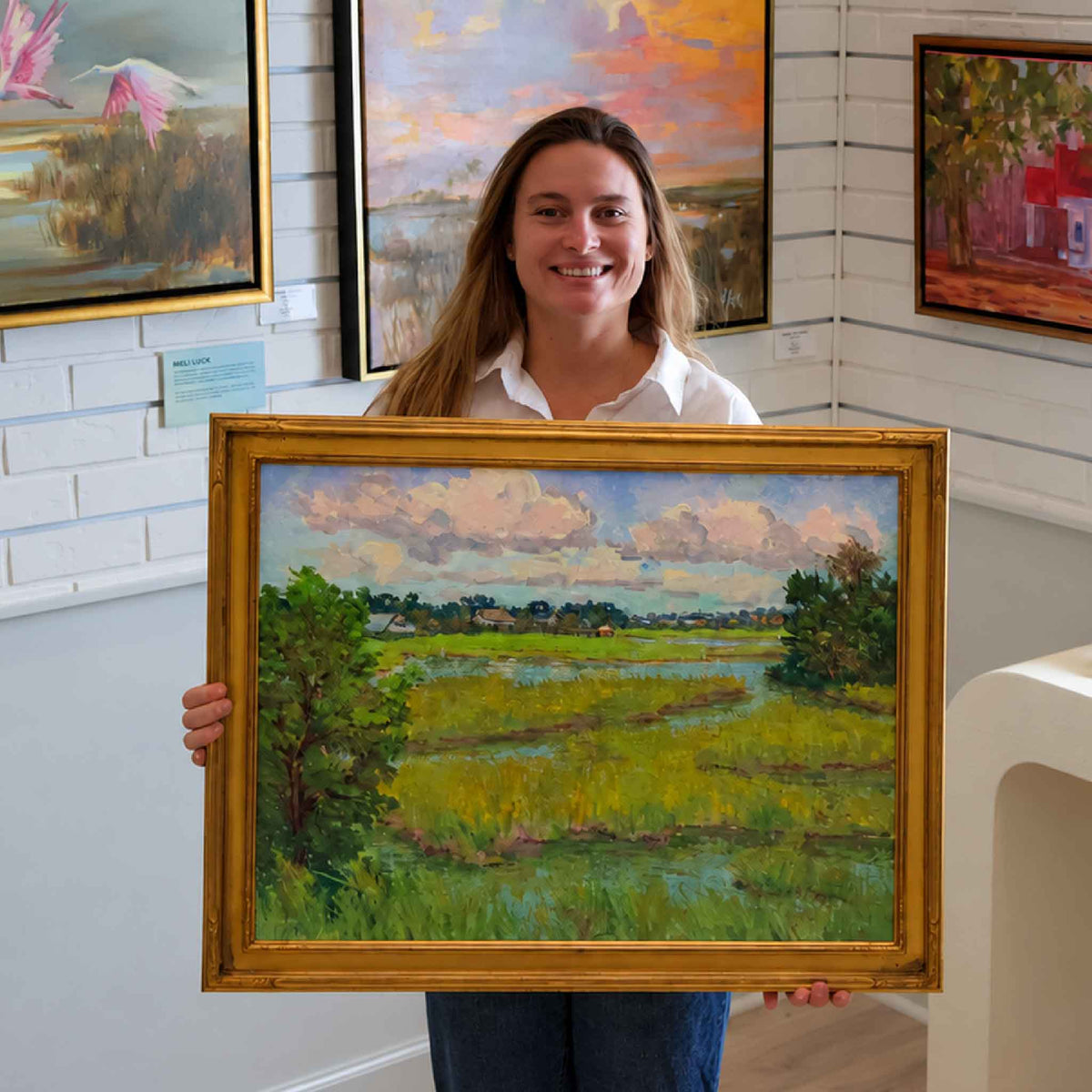 Gray Man Gallery team member holding Early Fall Marsh, an original oil painting by local artist Michael Rogan, showing the scale of the framed Lowcountry marsh landscape inside the Pawleys Island gallery.