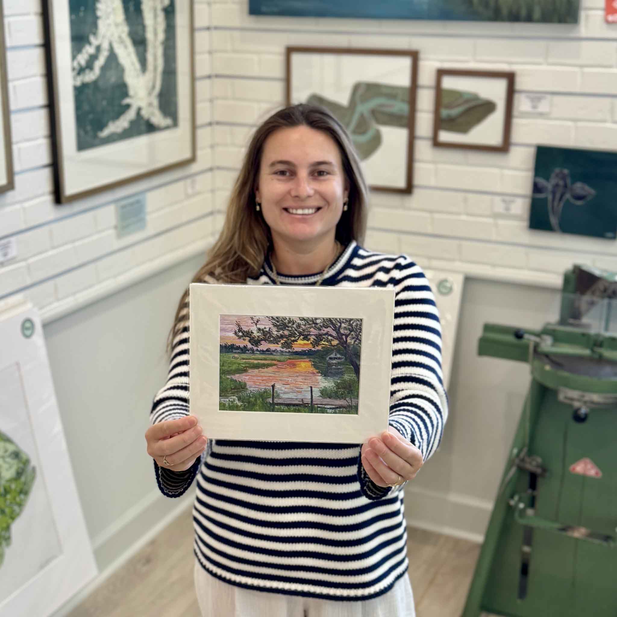 Kyra displays the 8×10 matted Fading Light fine art giclée print by Michael Rogan at Gray Man Gallery in Pawleys Island, featuring a South Litchfield marsh, tidal creek, dock, and colorful Lowcountry sunset.