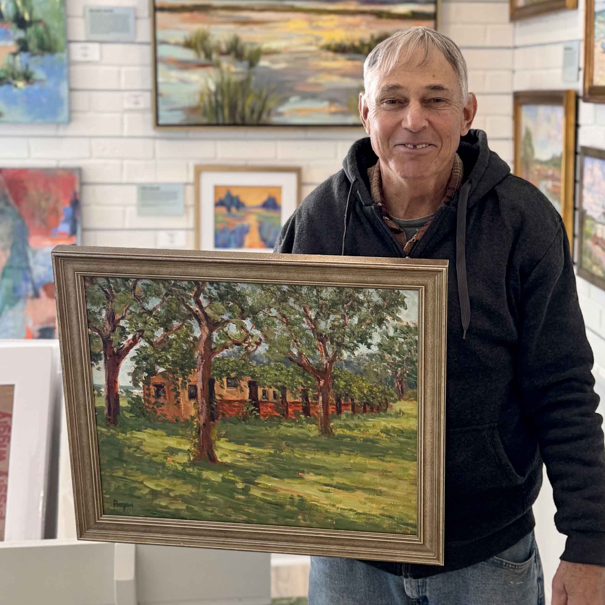 Michael Rogan, local artist, holding his original oil painting McKenzie Beach Oaks inside Gray Man Gallery, showcasing a Lowcountry oak-lined landscape inspired by McKenzie Beach in Pawleys Island, South Carolina
