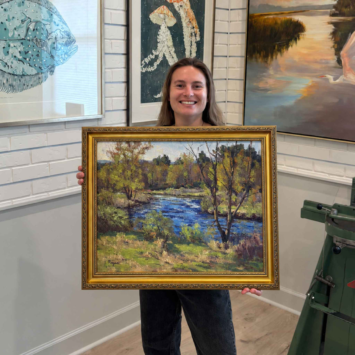 Framed Michael Rogan original oil painting Quiet Bend of the River displayed at Gray Man Gallery in Pawleys Island, featuring a vivid blue river surrounded by trees, grasses and layered green and golden foliage in an ornate gold frame