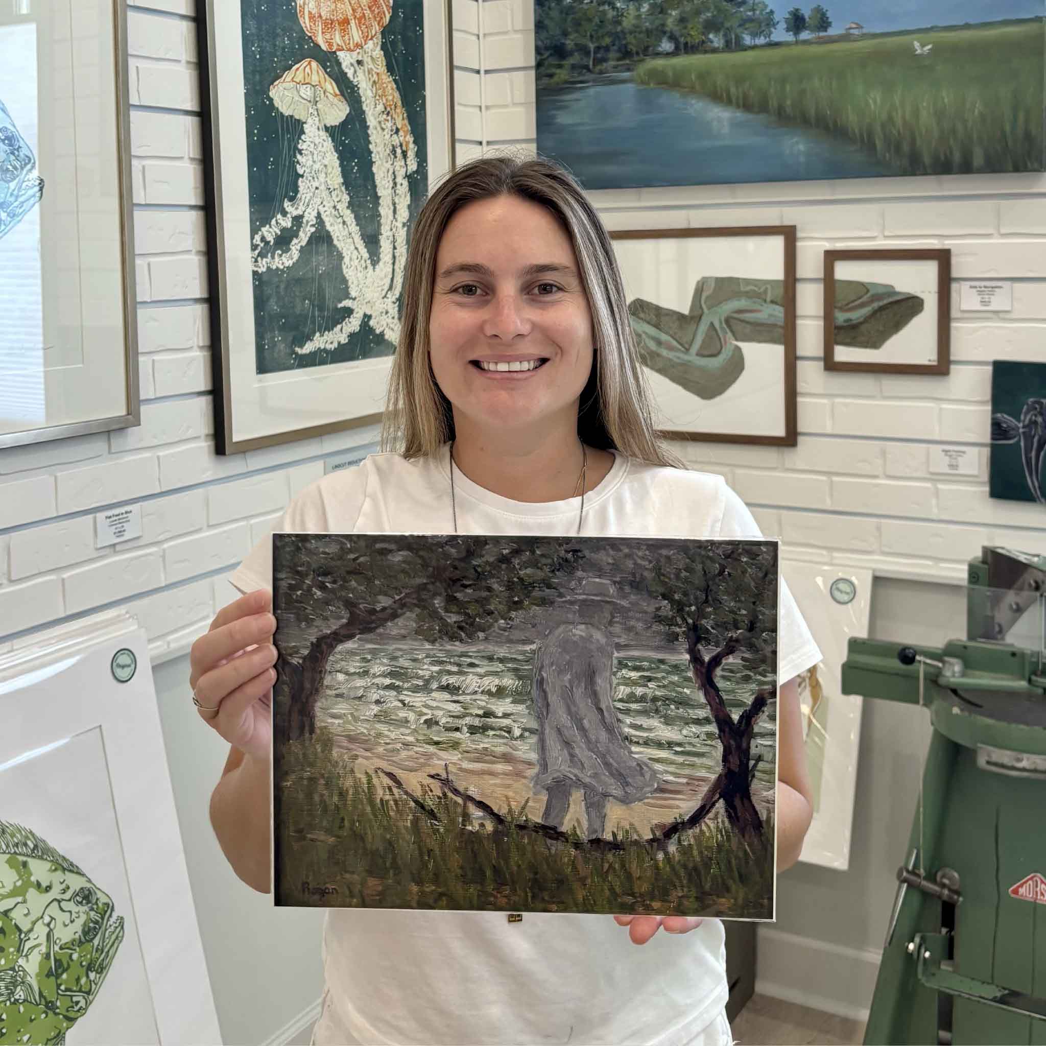 Kyra holds The Gray Man fine art giclée print by Lowcountry artist Michael Rogan at Gray Man Gallery in Pawleys Island, South Carolina, featuring Rogan’s interpretation of the legendary Gray Man walking along the windswept shoreline.