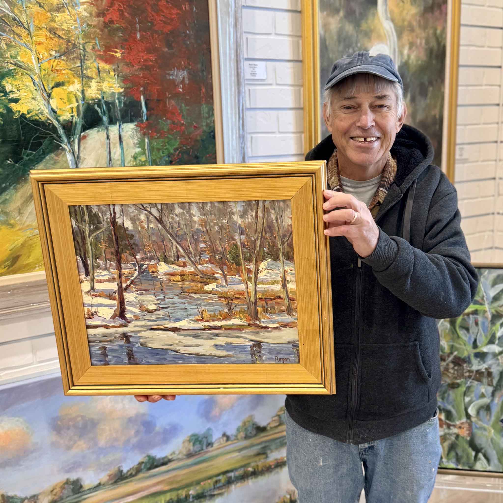 Artist Michael Rogan holding his framed Winter Creek Crossing original oil painting  inside Gray Man Gallery with artwork in the background.