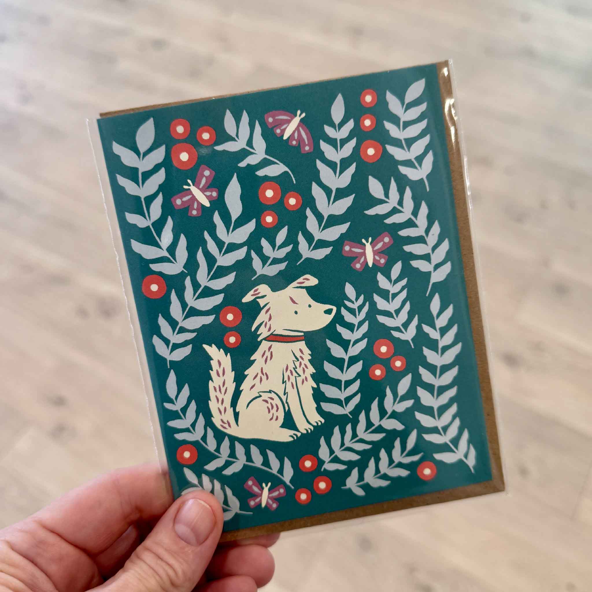 Customer holding the Molly Meadows Dog Greeting Card by Nuthatch Studio inside Gray Man Gallery in Pawleys Island, South Carolina. This whimsical botanical greeting card features a cream-colored dog surrounded by leafy green branches, bright coral berries, and fluttering butterflies on a rich teal background. Printed on premium 100% recycled paper and paired with a recycled kraft envelope, it's a charming eco-friendly greeting card for birthdays, thank you notes, encouragement, or everyday correspondence.