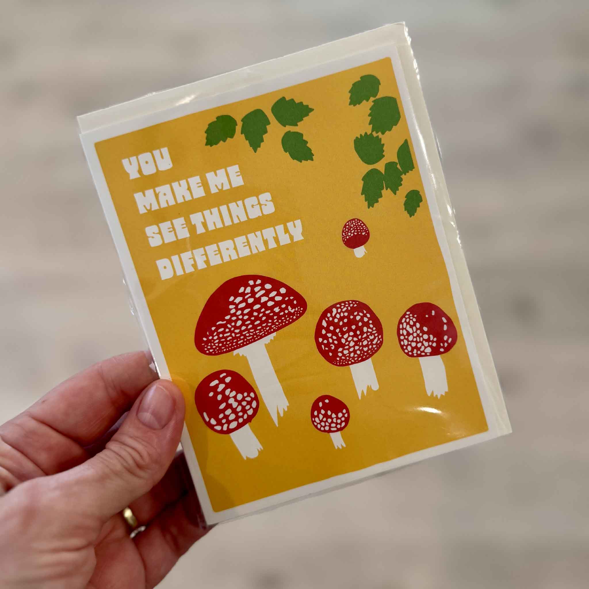 You Make Me See Things Differently mushroom greeting card by Lonely Palm Ranch held inside Gray Man Gallery in Pawleys Island, with red and white mushrooms, green leaves and bold retro lettering on a golden yellow background.