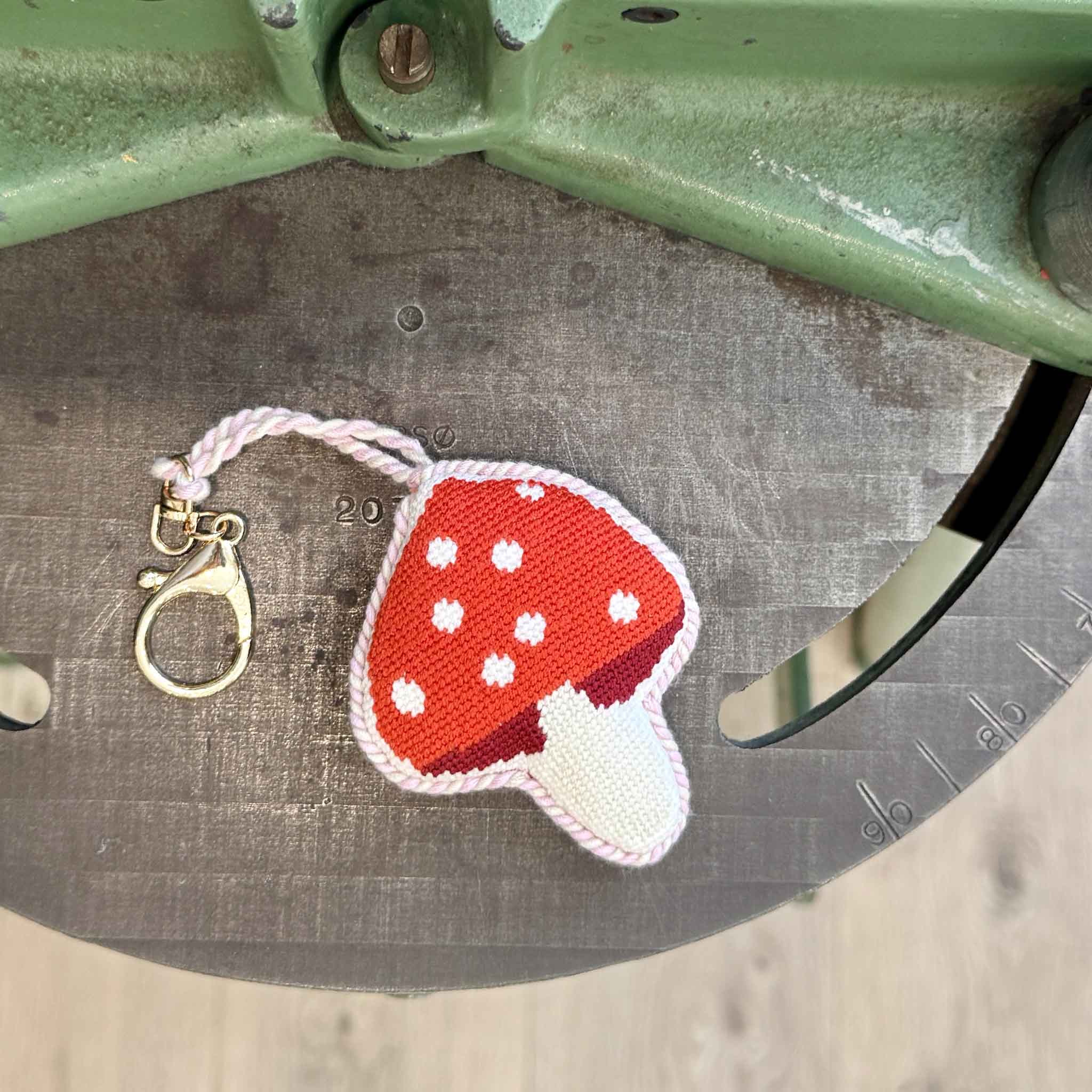 Hand-stitched mushroom needlepoint bag charm with a red-and-white toadstool design displayed atop vintage framing equipment inside the custom frame shop at Gray Man Gallery in Pawleys Island, South Carolina. Featuring detailed needlepoint embroidery, a soft velvet backing, twisted pink cord, and gold-tone lobster clasp, this whimsical accessory easily clips onto handbags, backpacks, travel totes, key rings, or luggage for a fun woodland-inspired accent.