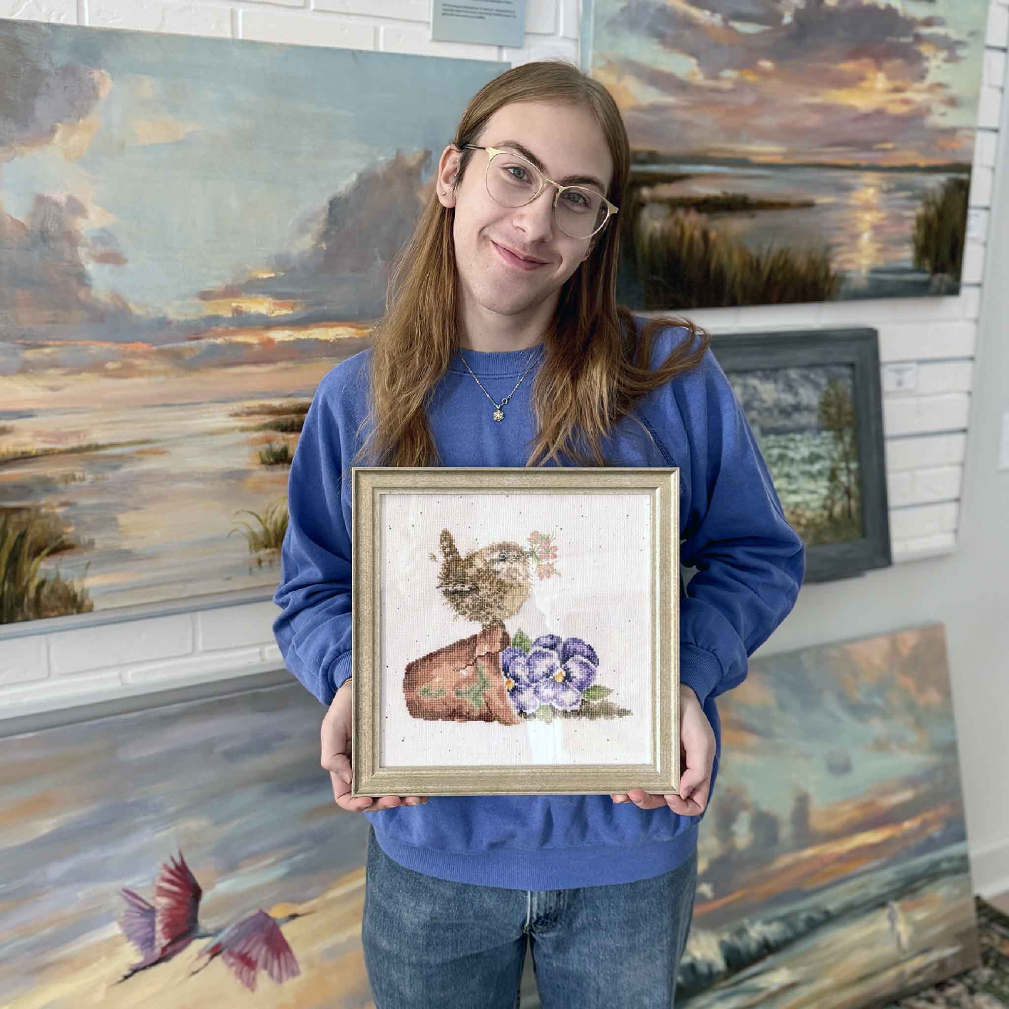 A Gray Man Gallery team member holds Nestled in the Garden by Mimi Beaver, a framed cross-stitch featuring a small bird perched beside flowers, donated to benefit Salty Dogs Senior Animal Rescue.