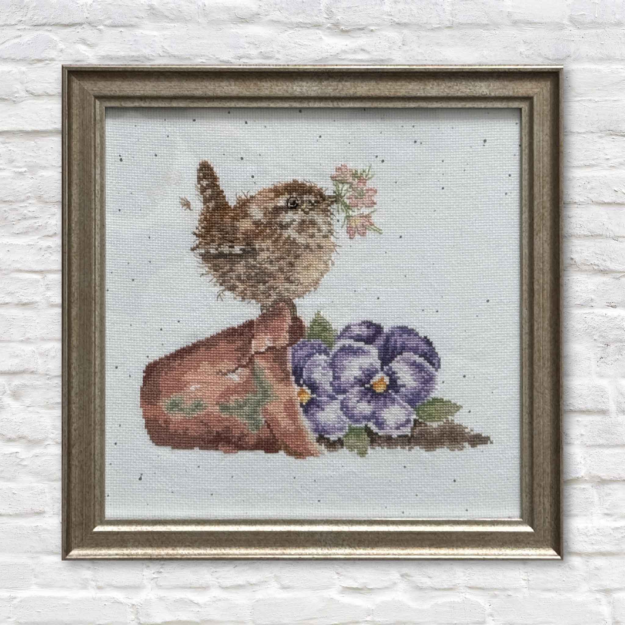 Framed cross-stitch artwork titled Nestled in the Garden by Mimi Beaver, featuring a small bird with flowers in a garden scene, donated to support senior dog rescue.