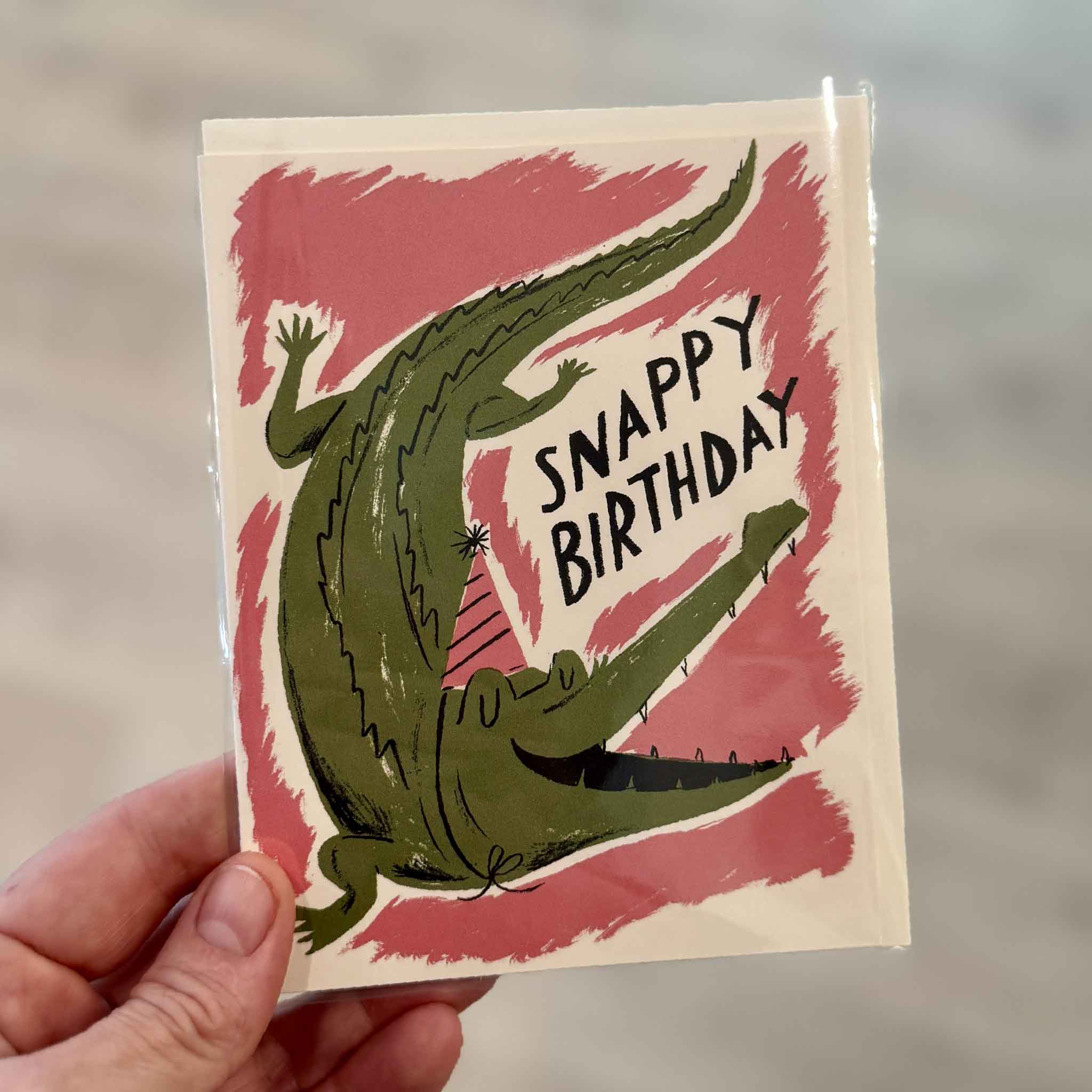 Snappy Birthday greeting card illustrated by Nicholas John Frith for Red Cap Cards, photographed inside Gray Man Gallery in Pawleys Island, South Carolina. This humorous birthday card features a smiling alligator wearing a party hat with the playful phrase "Snappy Birthday."