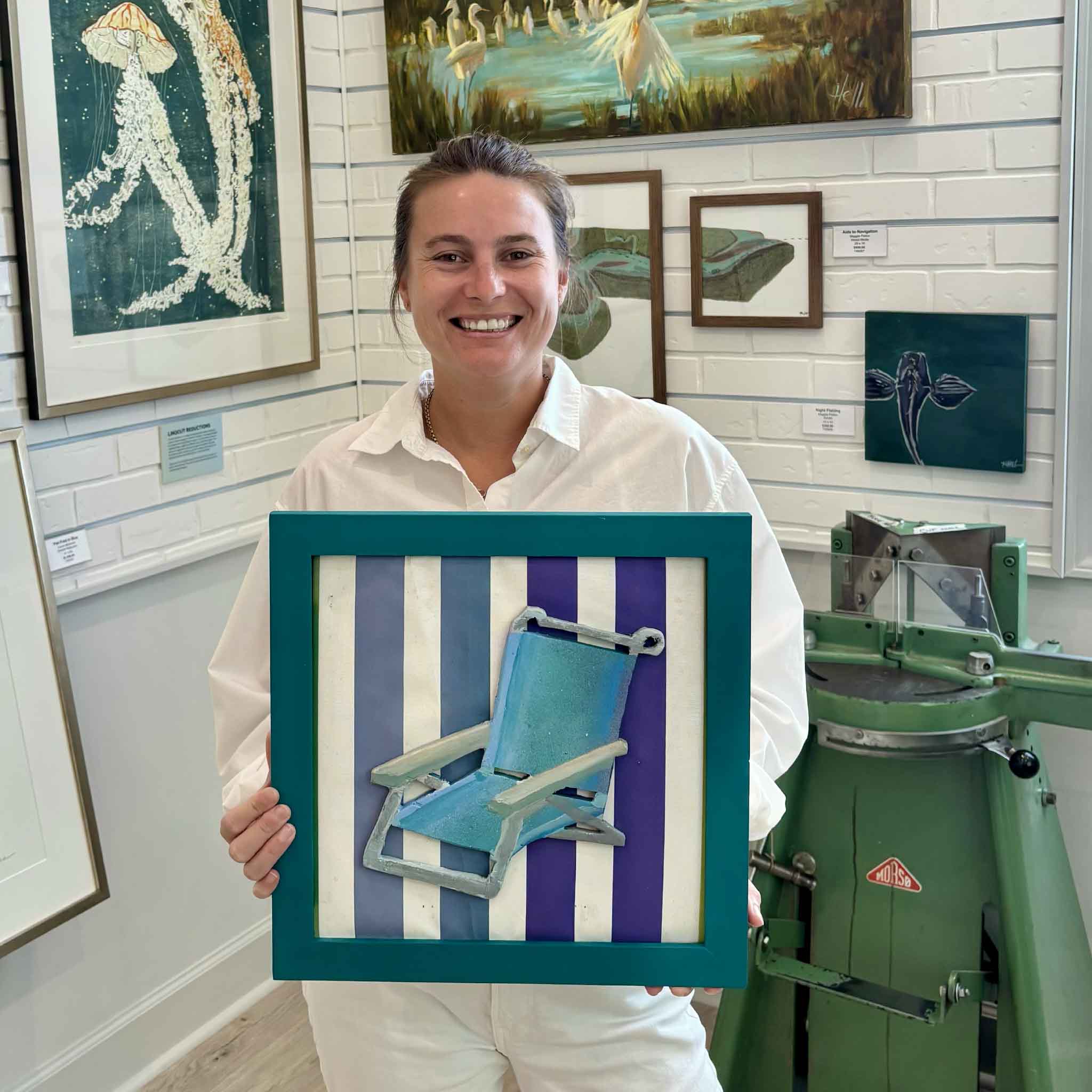 Kyra holding Beach Chair by Charleston artist Nick Kawczynski, a one-of-a-kind original coastal assemblage with a miniature chair constructed from found wood and a striped background repurposed from an old beach chair, inside Gray Man Gallery in Pawleys Island.