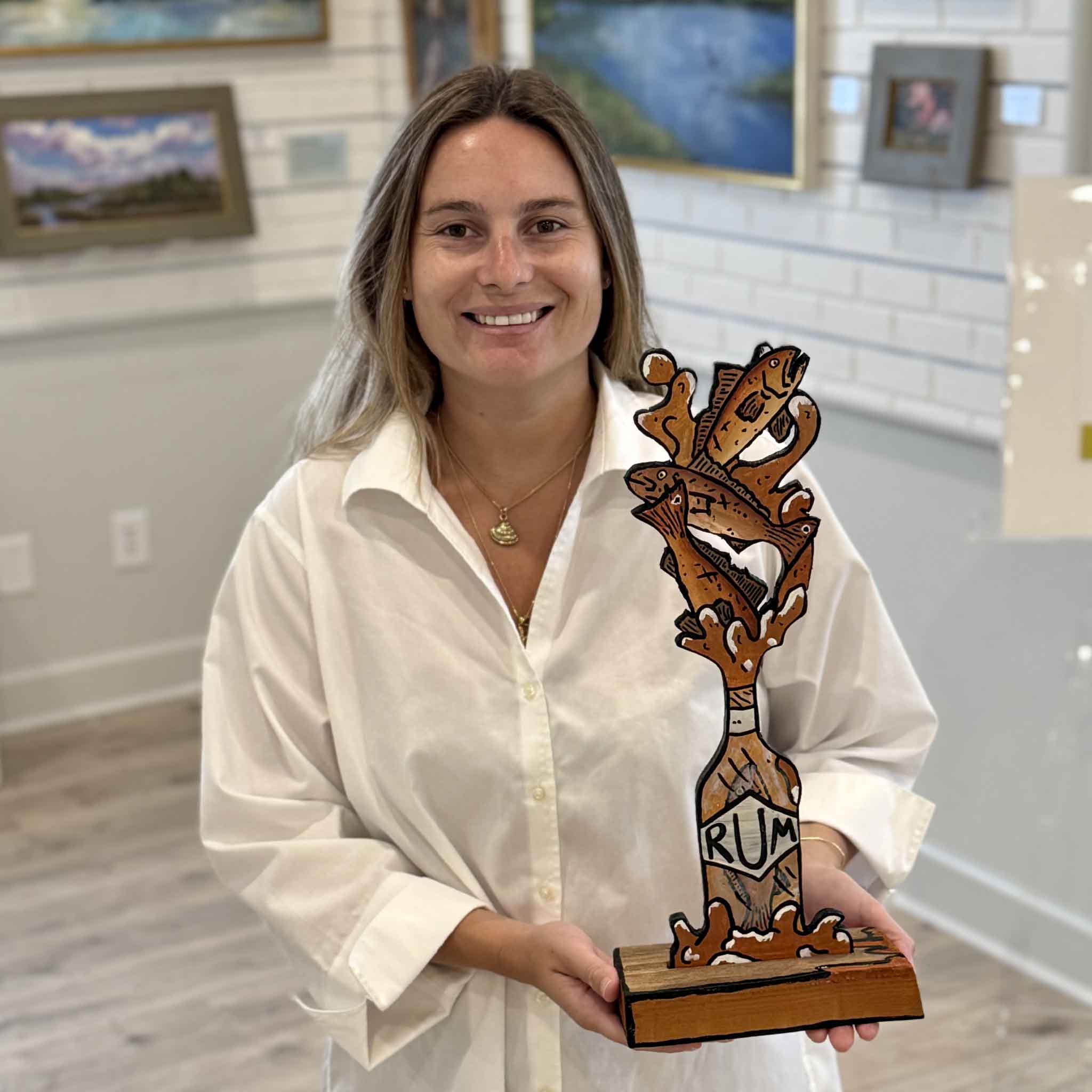 Kyra holds Drum 'n Rum, an original mixed media sculpture by Charleston artist Nick Kawczynski. This handcrafted coastal artwork features red drum fish, a rum bottle motif, reclaimed marine-grade plywood, and a reclaimed wood base. Available exclusively through Gray Man Gallery in Pawleys Island, South Carolina.