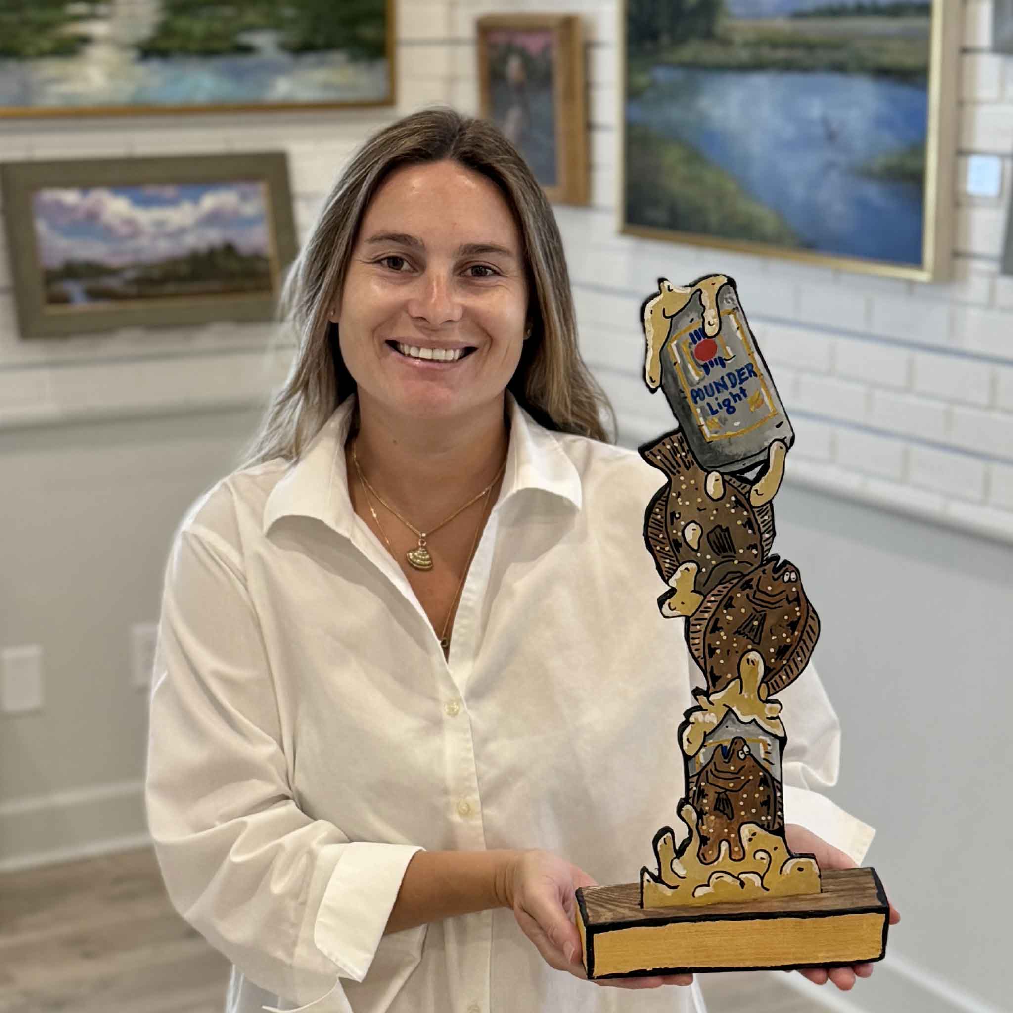 Kyra holds Poundahs 'n Floundahs, an original mixed media sculpture by Charleston artist Nick Kawczynski. Featuring three hand-painted flounder, a playful Pounder Light beer can motif, reclaimed marine-grade plywood, and a handcrafted wood base, this one-of-a-kind sculpture is available exclusively at Gray Man Gallery in Pawleys Island, South Carolina.