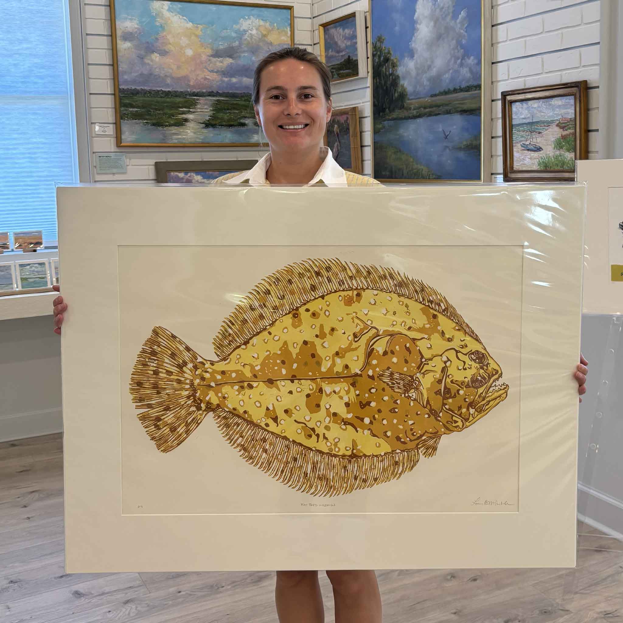 Kyra holds Flat Fred in Lemon, an original signed reduction linocut by Laurie Brownell McIntosh featuring a golden yellow flounder in layered ochre and amber inks. Professionally matted to fit a standard 30x40 frame, this iconic Lowcountry print is available at Gray Man Gallery in Pawleys Island, South Carolina.