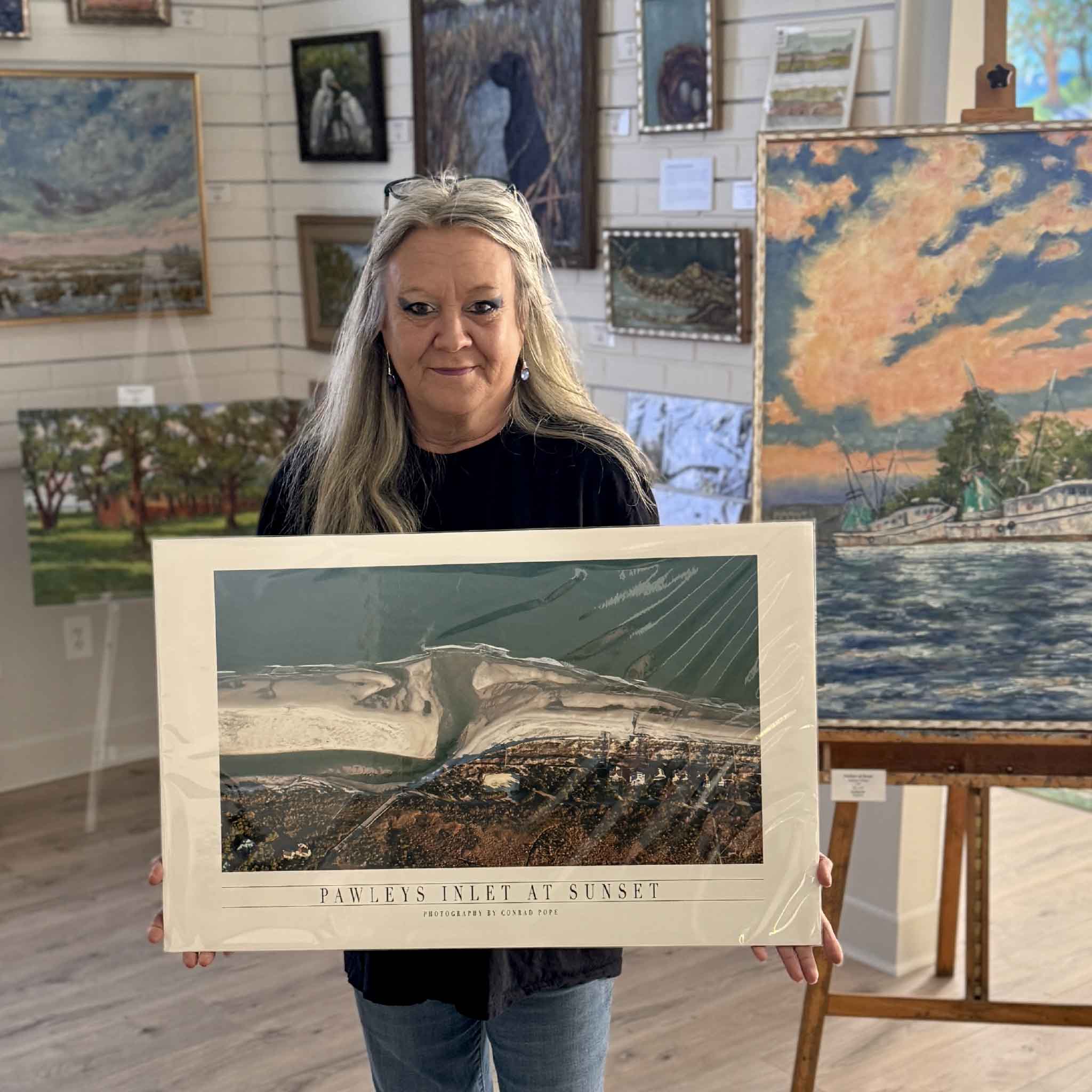 Charlotte holding local photographer Conrad Pope's  Pawleys Inlet at Sunset aerial photograph inside Gray Man Gallery, showcasing a large coastal print that captures the natural beauty of Pawleys Island at sunset.