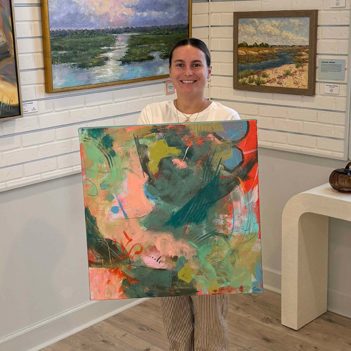 Gray Man Gallery team member holding Peach Blossom, a colorful original gallery-wrapped acrylic painting by local artist Susan Moore, inside the gallery in Pawleys Island, South Carolina. The large contemporary abstract artwork features sweeping teal brushstrokes, bright coral and peach accents, soft green passages, and layered painterly textures, with the photograph showing its scale and unframed canvas presentation.