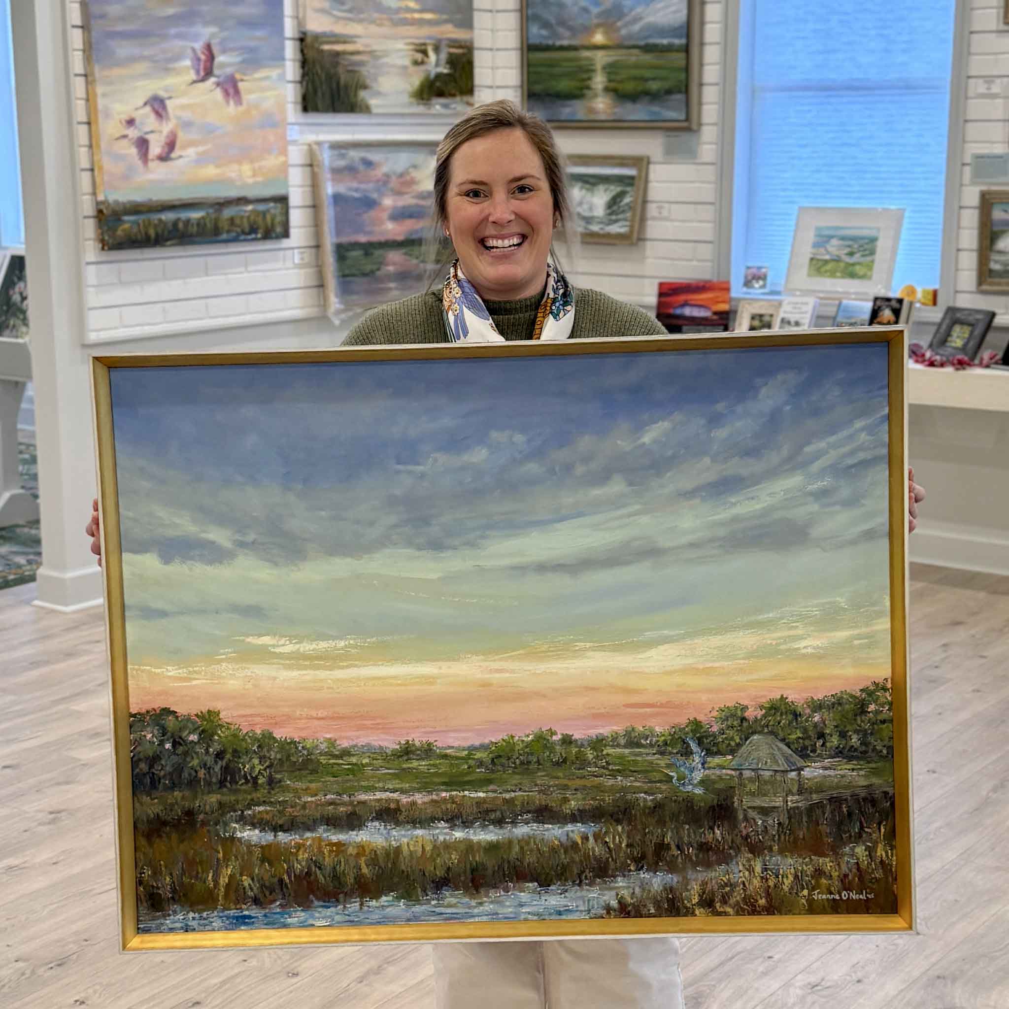 Bordy holding Peach Moonshine, an original oil painting by local Lowcountry artist Jeanne O’Neal, showing framed marsh sunset landscape with heron and pastel sky inside Gray Man Gallery in Pawleys Island, South Carolina