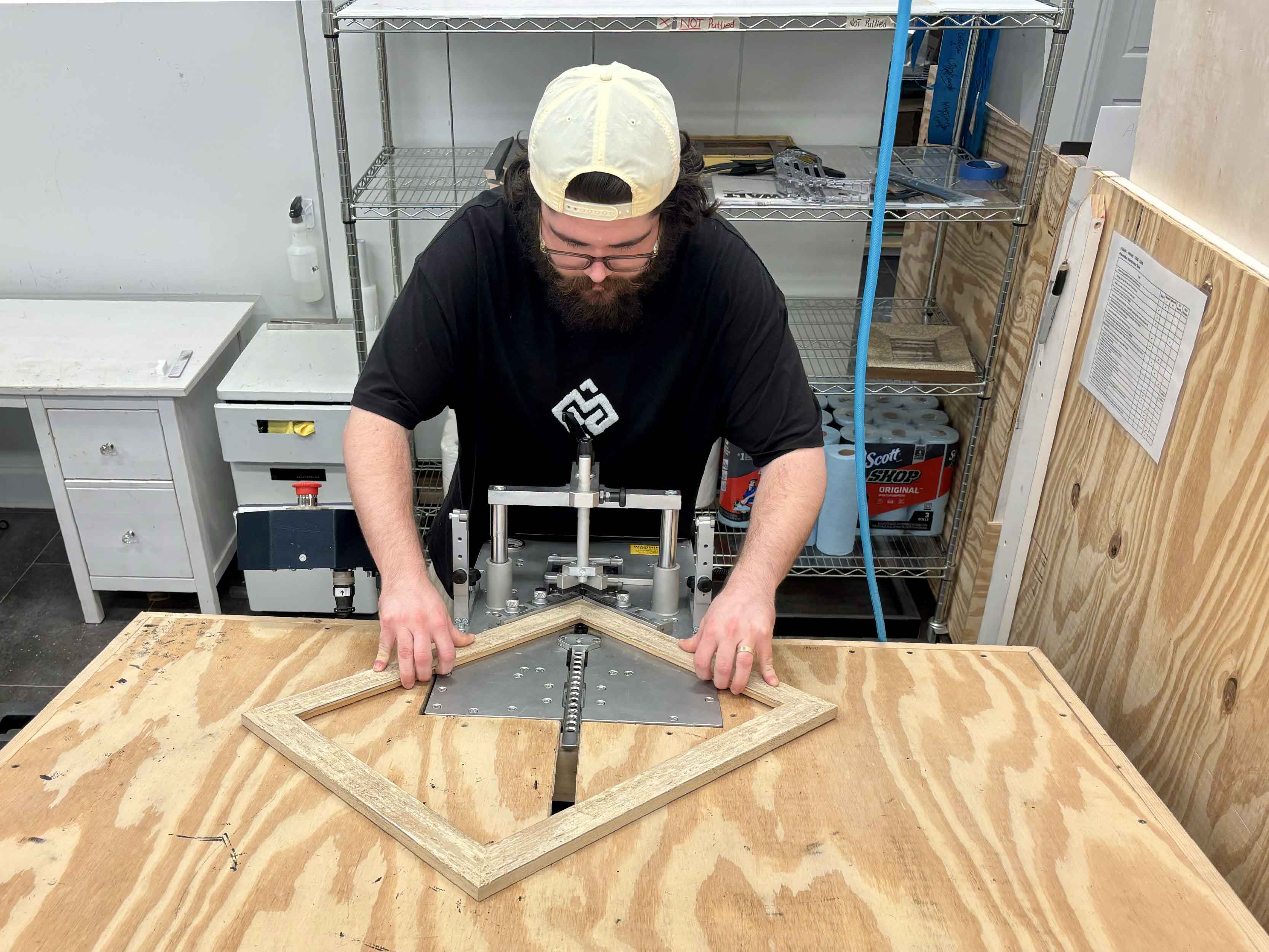 Gray Man Gallery custom framer Peyton carefully joins and assembles a custom picture frame by hand in the professional frame shop at Gray Man Gallery in Pawleys Island, South Carolina. The image shows the precision frame-joining process used to align and secure each corner before artwork, matting, conservation glass, backing, and finishing hardware are assembled into the completed custom frame.