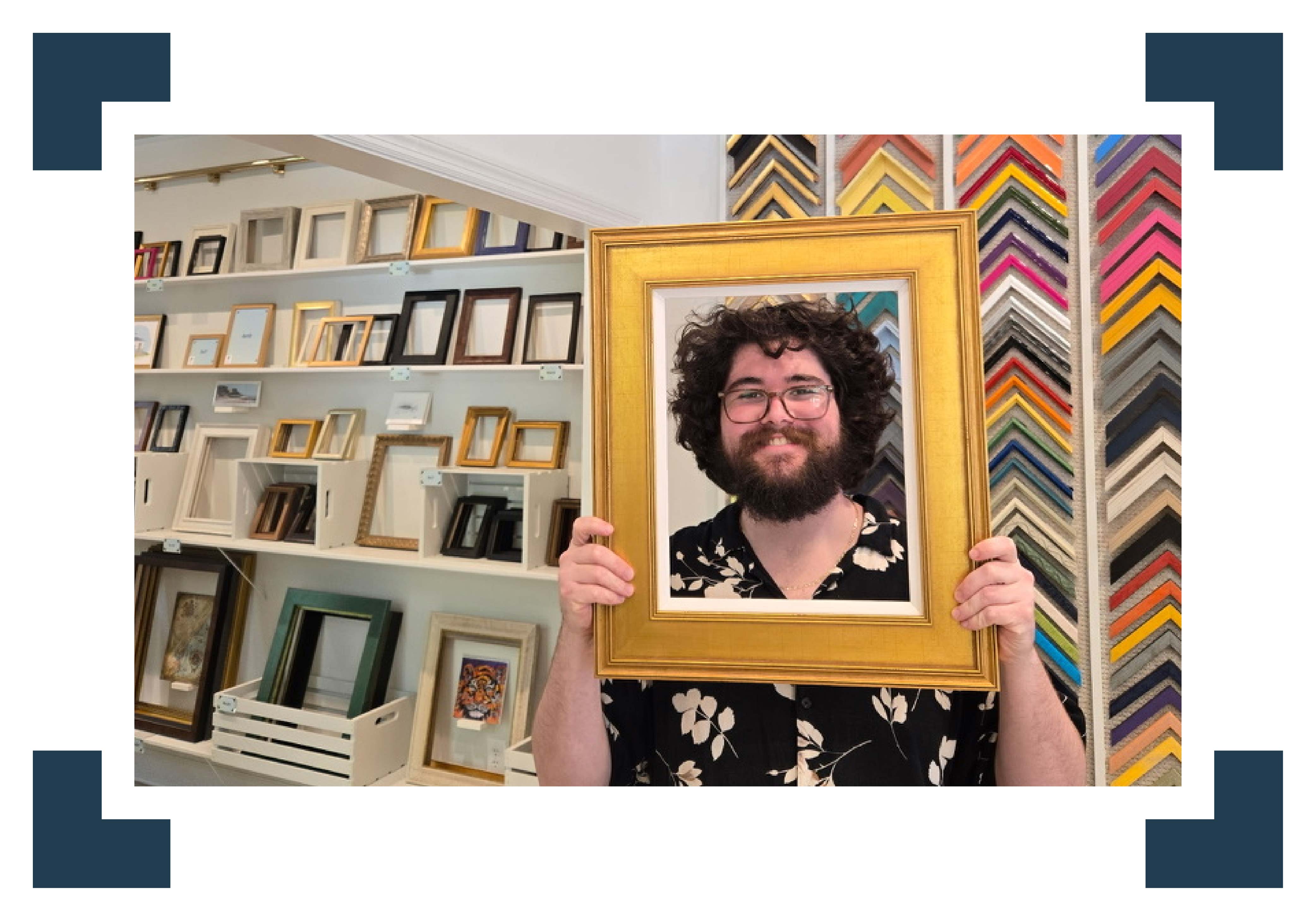 Peyton smiles while holding a classic gold picture frame up like a portrait at Gray Man Gallery in Pawleys Island, South Carolina, standing in front of walls filled with colorful frame corner samples and ready-made frames, showcasing the gallery’s wide selection of custom framing styles and the approachable, boutique Lowcountry art and framing experience