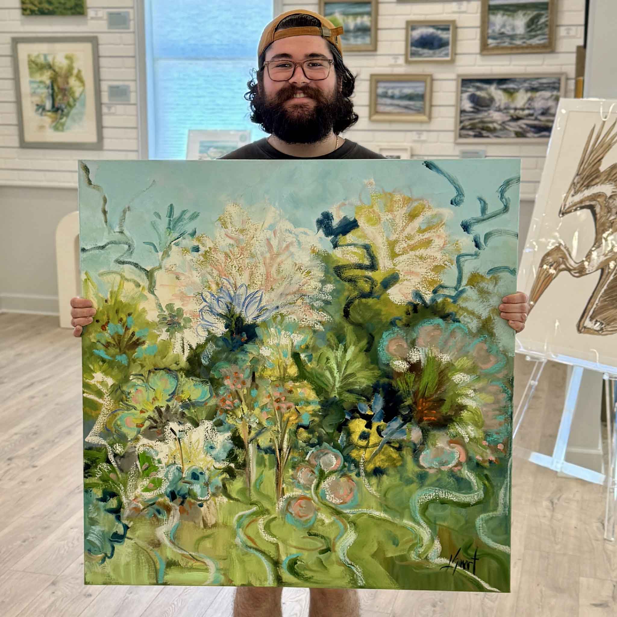 Peyton holds Glorious Sea Fans, a 36 × 36 original gallery-wrapped oil painting by Libby Smart, inside Gray Man Gallery in Pawleys Island, South Carolina. The photograph showcases the scale of this contemporary coastal artwork featuring layered sea fans, blooming botanicals, flowing organic forms, and a palette of seafoam green, turquoise blue, coral, and white inspired by the beauty of the Lowcountry.