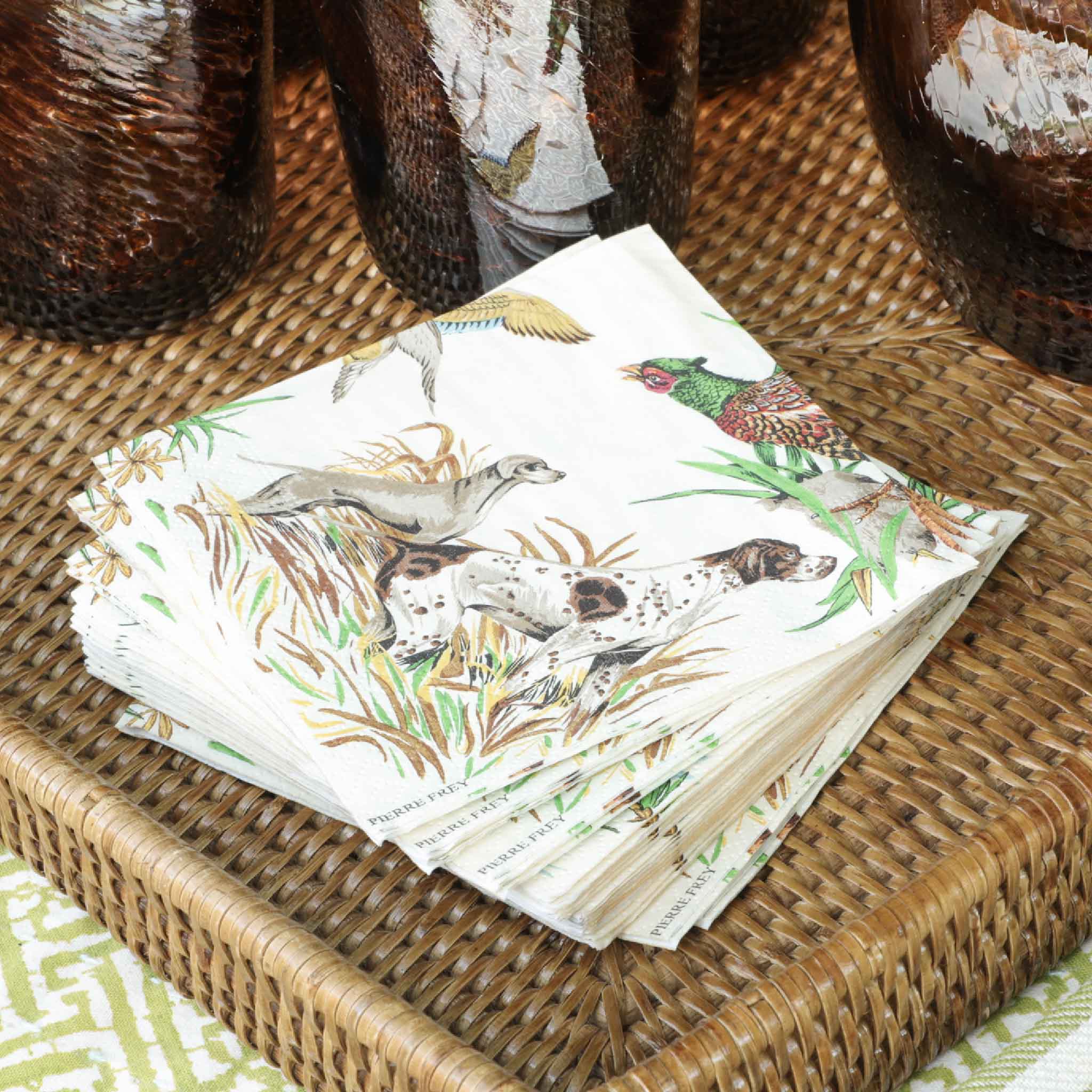 Pierre Frey Sologne Cocktail Napkins by Caspari displayed on a woven rattan serving tray alongside textured amber glassware. The elegant duck and marsh design creates a refined table setting for cocktail parties, sporting lodges, lake houses, coastal entertaining, and fall gatherings. Available at Gray Man Gallery in Pawleys Island, South Carolina.