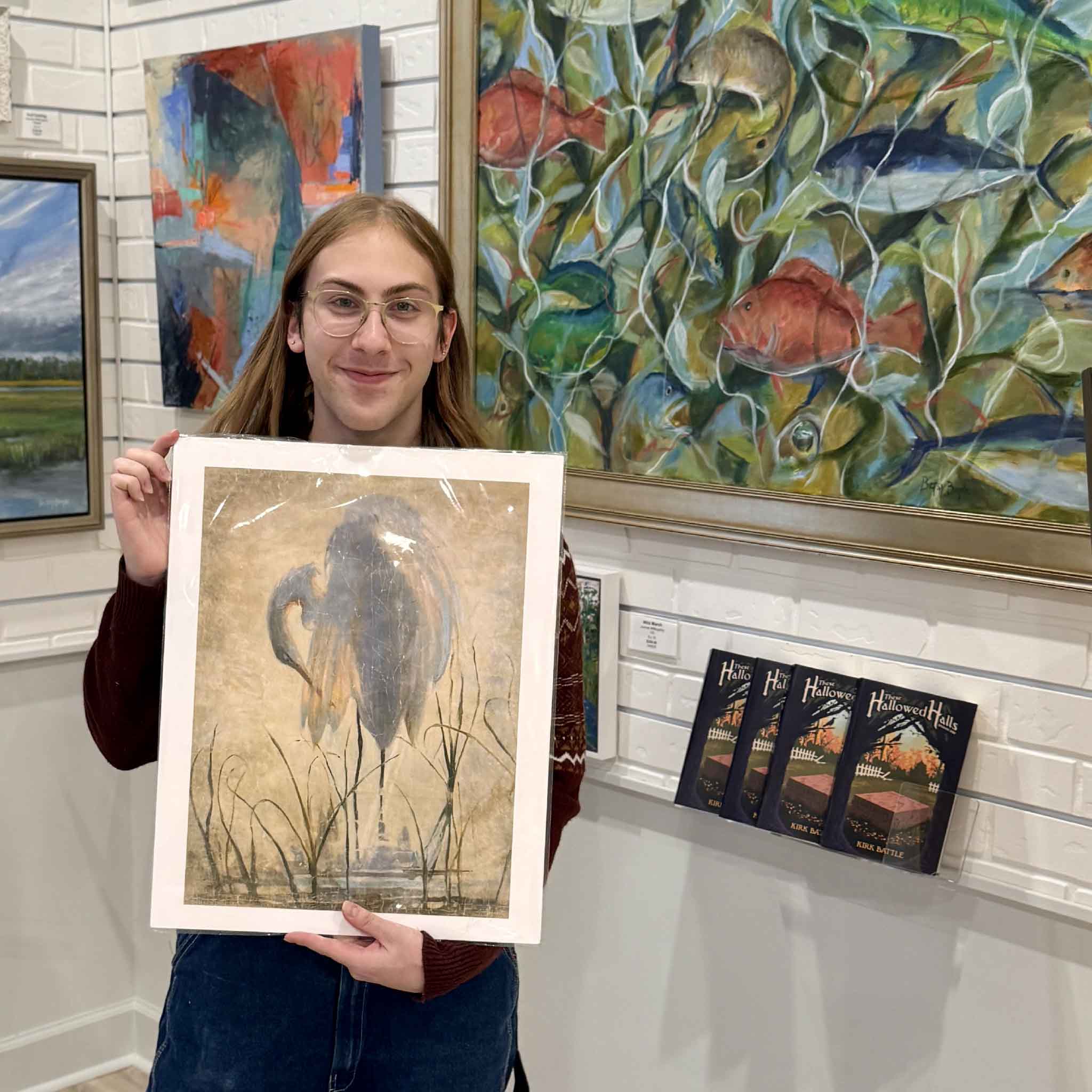 A gallery assistant holds the 16x20Preening Egret art print by Helli Luck, showing the scale and elegant detail of the Lowcountry bird illustration against a backdrop of original coastal artwork at Gray Man Gallery.