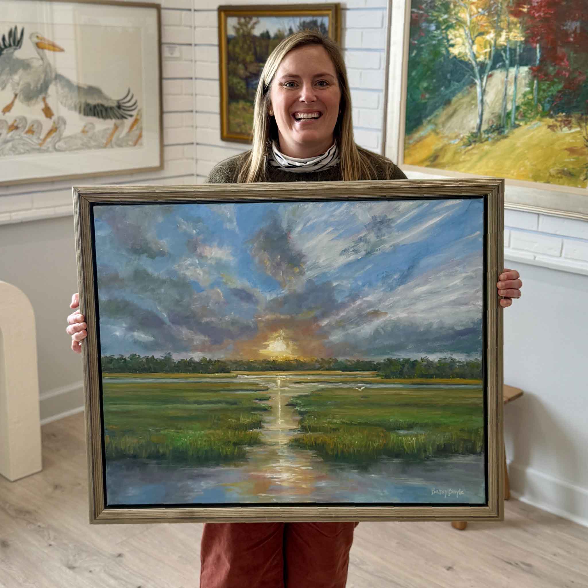 Gray Man Gallery team member holding Promise, a 30x24 original oil painting by local South Carolina artist Betsy Boyle, photographed inside the Pawleys Island art gallery with custom framing and Lowcountry artwork on display
