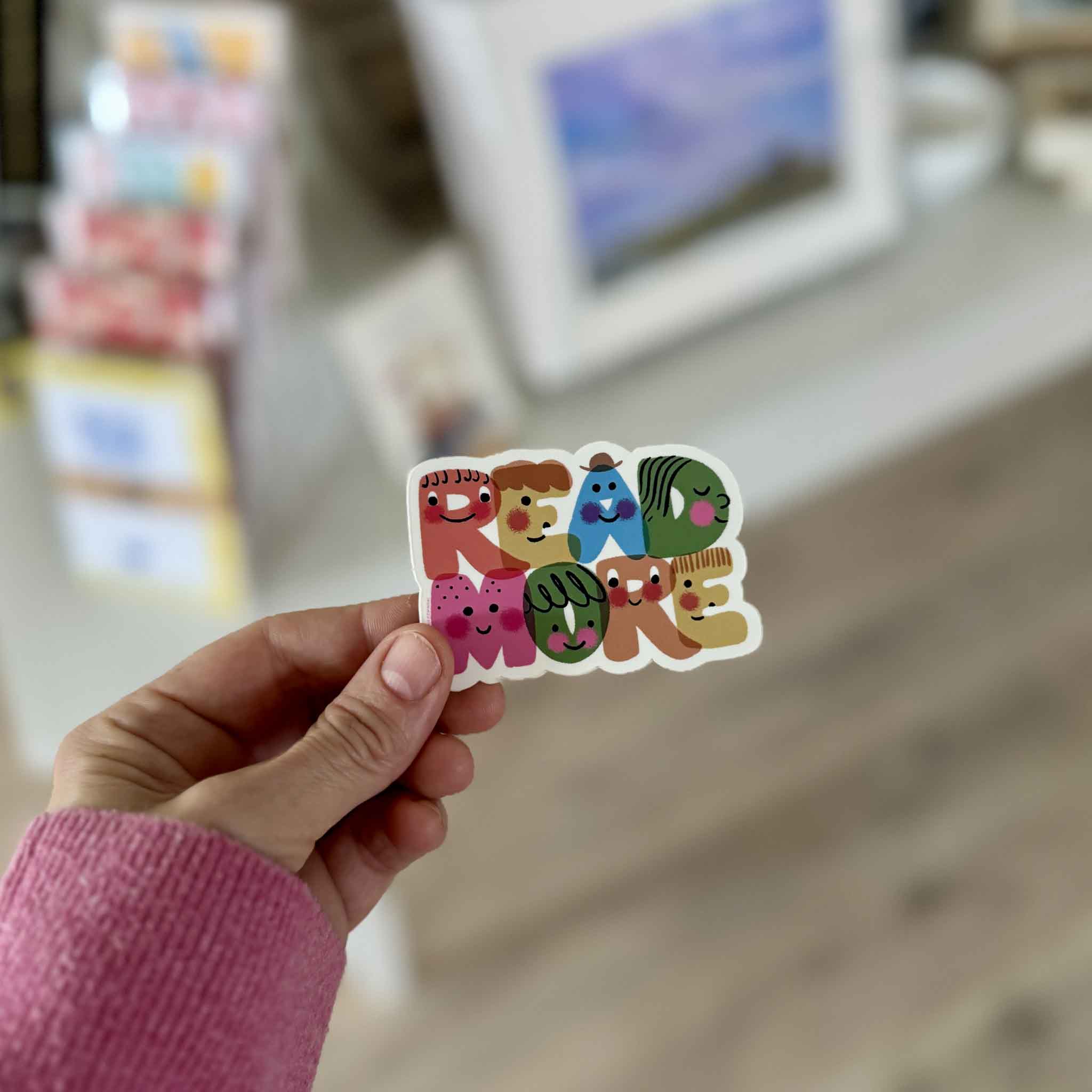 Hand holding the colorful Read More vinyl sticker by illustrator Dylan Mierzwinski inside Gray Man Gallery in Pawleys Island, showing the sticker’s playful smiling letter characters and compact size.