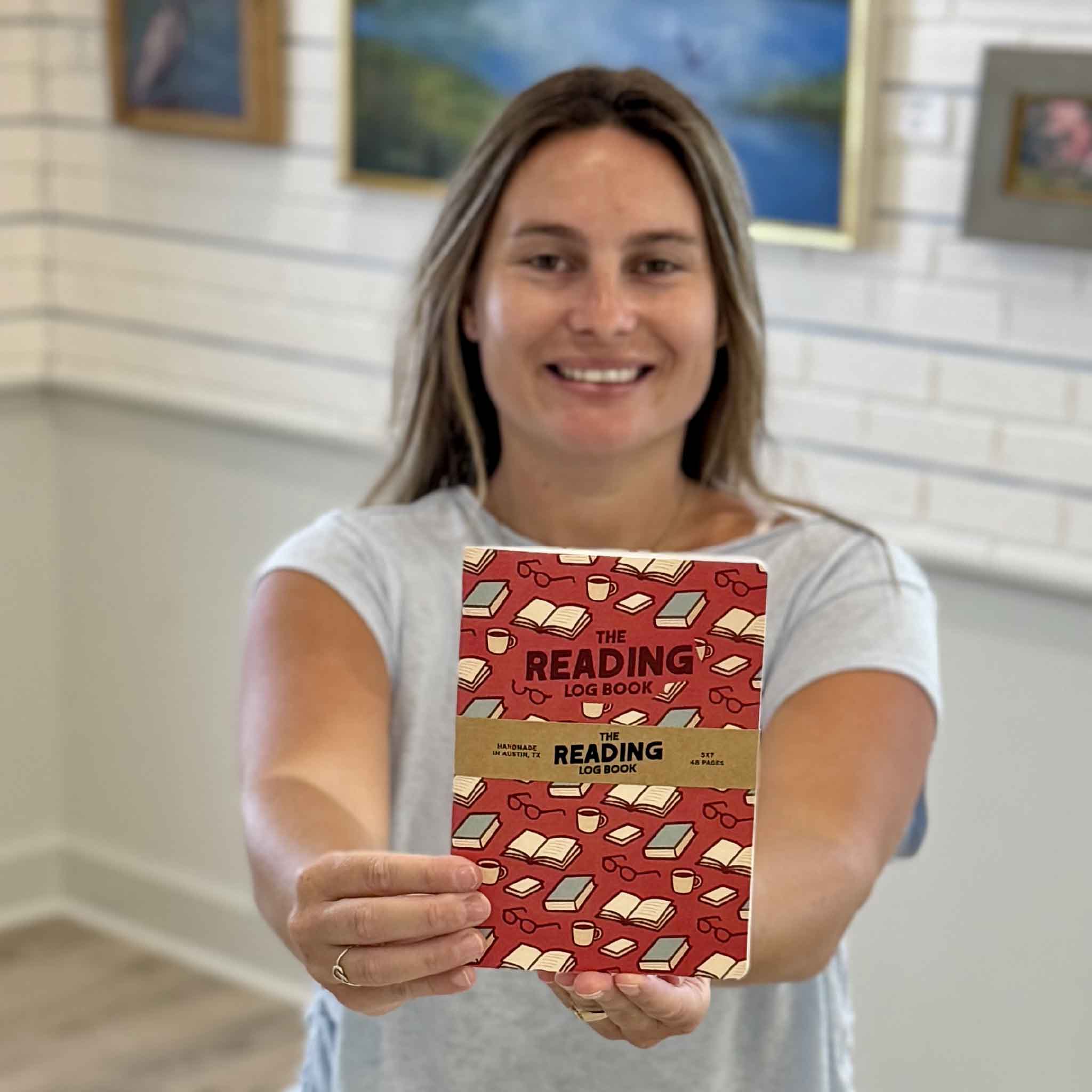 Gray Man Gallery team member Kyra holding The Reading Log Book inside the gallery. This 5x7 reading journal is designed for book lovers who want to record titles, authors, favorite quotes, ratings, reviews, and reflections from every book they read.