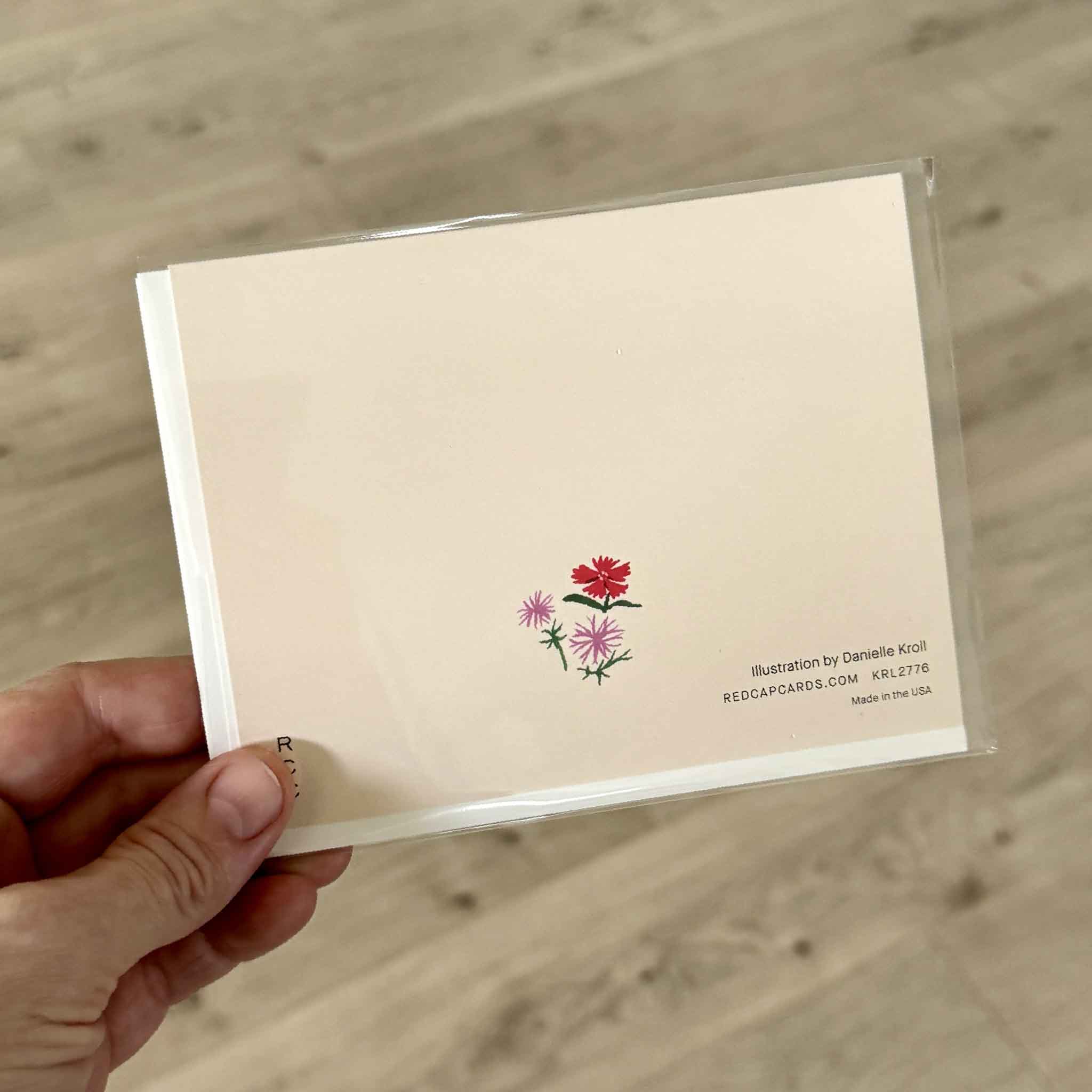 Back of the Red Cap Cards "Birthday Critters" greeting card illustrated by Danielle Kroll, photographed inside Gray Man Gallery in Pawleys Island, South Carolina. The back features the artist credit, floral artwork, and premium recycled cardstock.