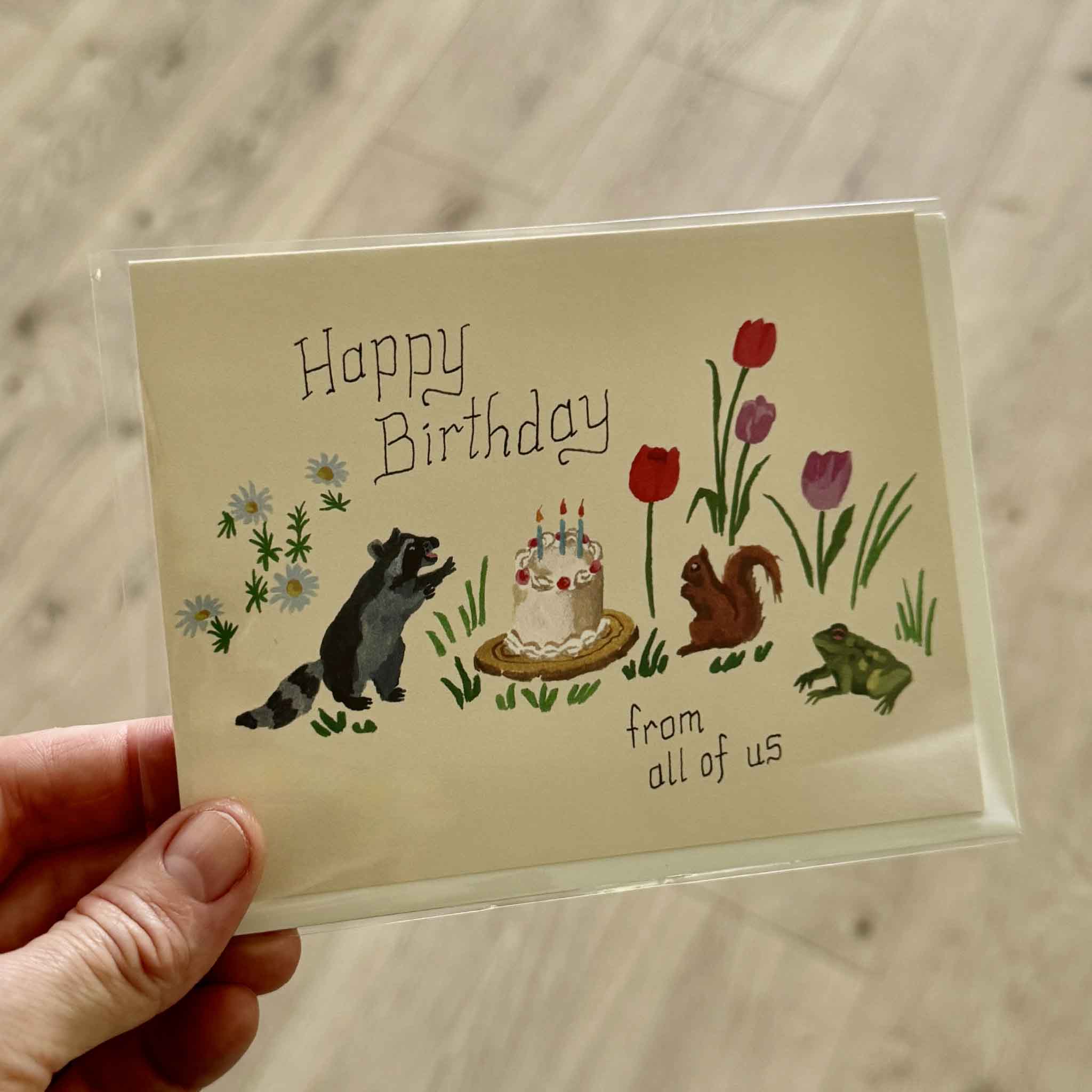 Hand holding the Red Cap Cards "Birthday Critters" greeting card illustrated by Danielle Kroll inside Gray Man Gallery in Pawleys Island, South Carolina. The whimsical birthday design features a raccoon, squirrel, frog, colorful tulips, daisies, and a birthday cake.