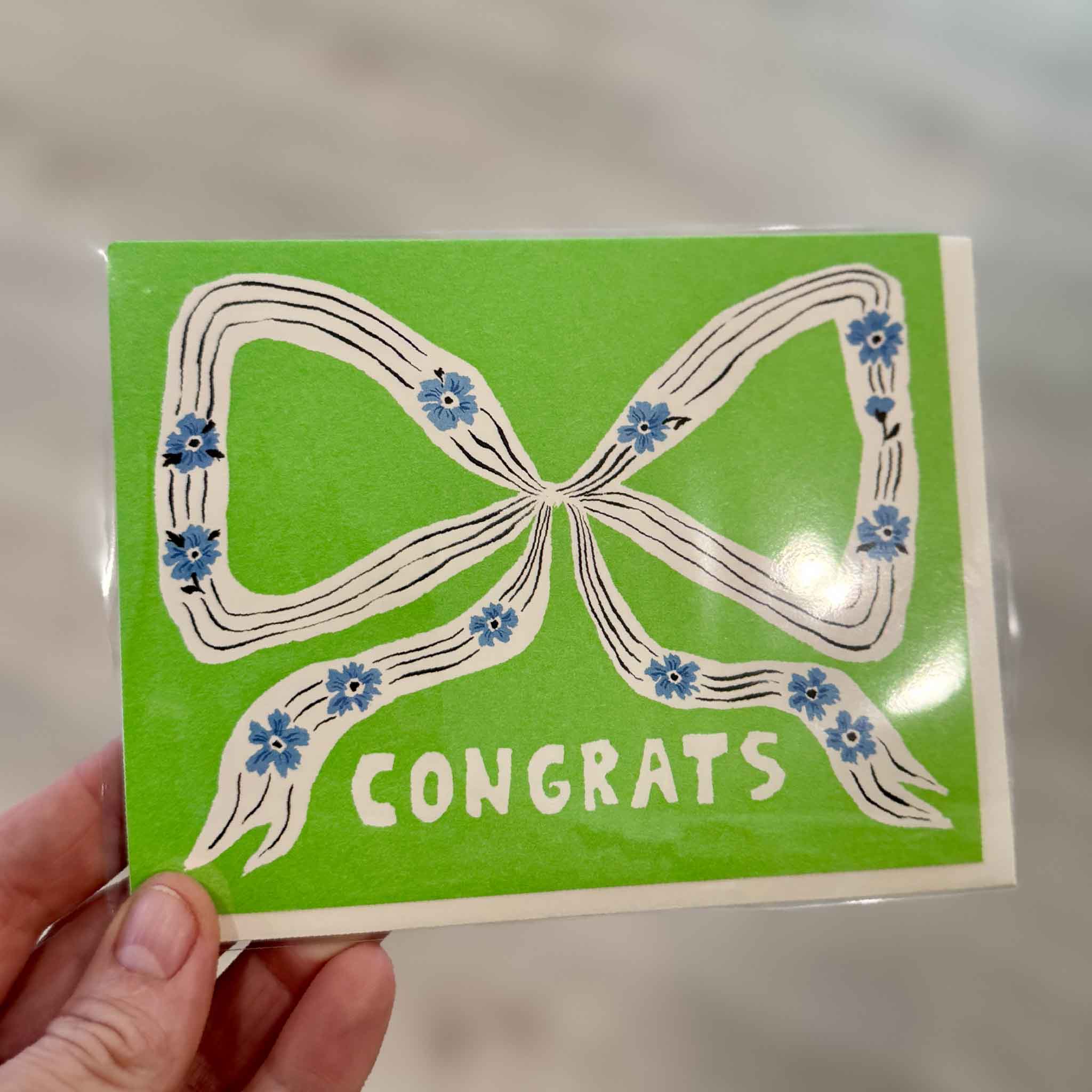 Perfect Bow congratulations greeting card by Emily Isabella from Red Cap Cards displayed inside Gray Man Gallery in Pawleys Island, South Carolina. This premium blank greeting card features a hand-painted floral ribbon and is ideal for graduations, weddings, promotions, new homes, retirements, and other celebratory occasions.
