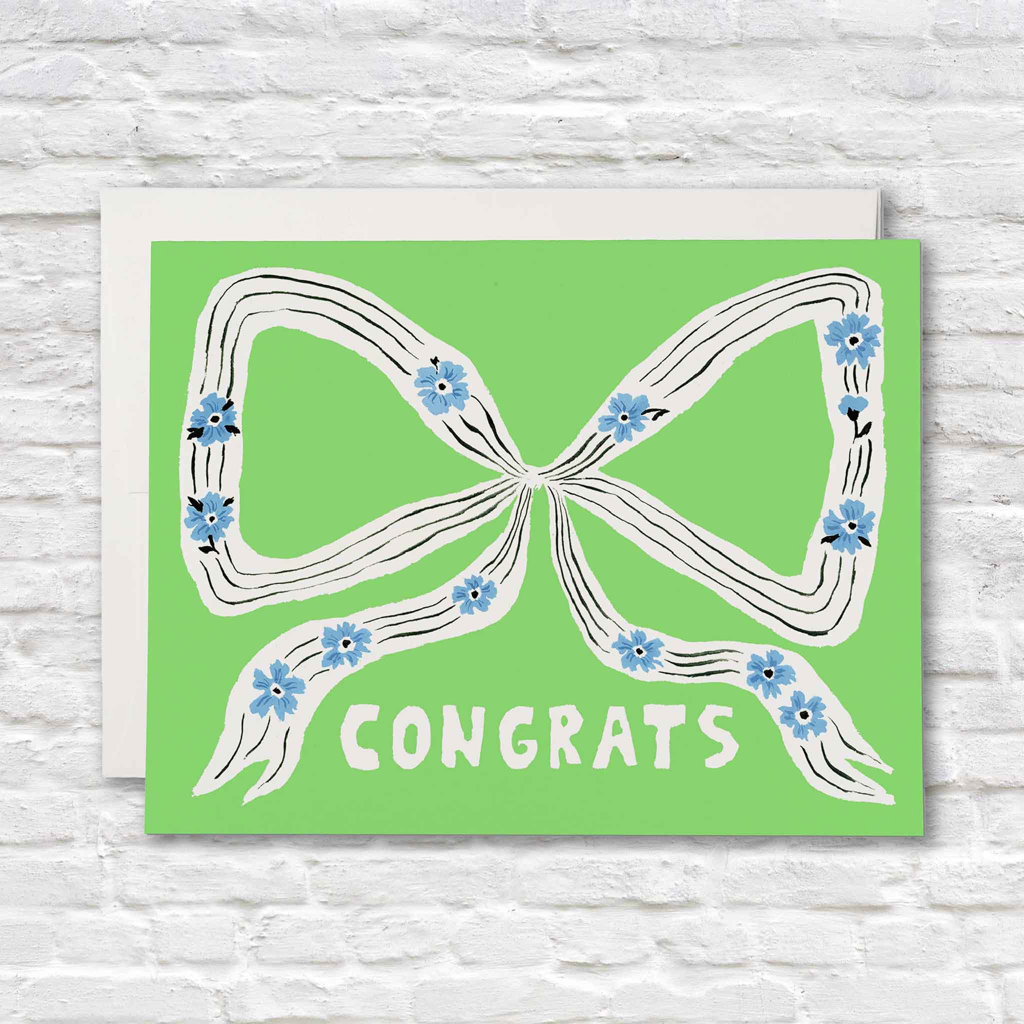 Perfect Bow congratulations greeting card illustrated by Emily Isabella for Red Cap Cards featuring a hand-painted white ribbon with blue flowers on a bright green background. Blank inside premium greeting card available at Gray Man Gallery in Pawleys Island, South Carolina.