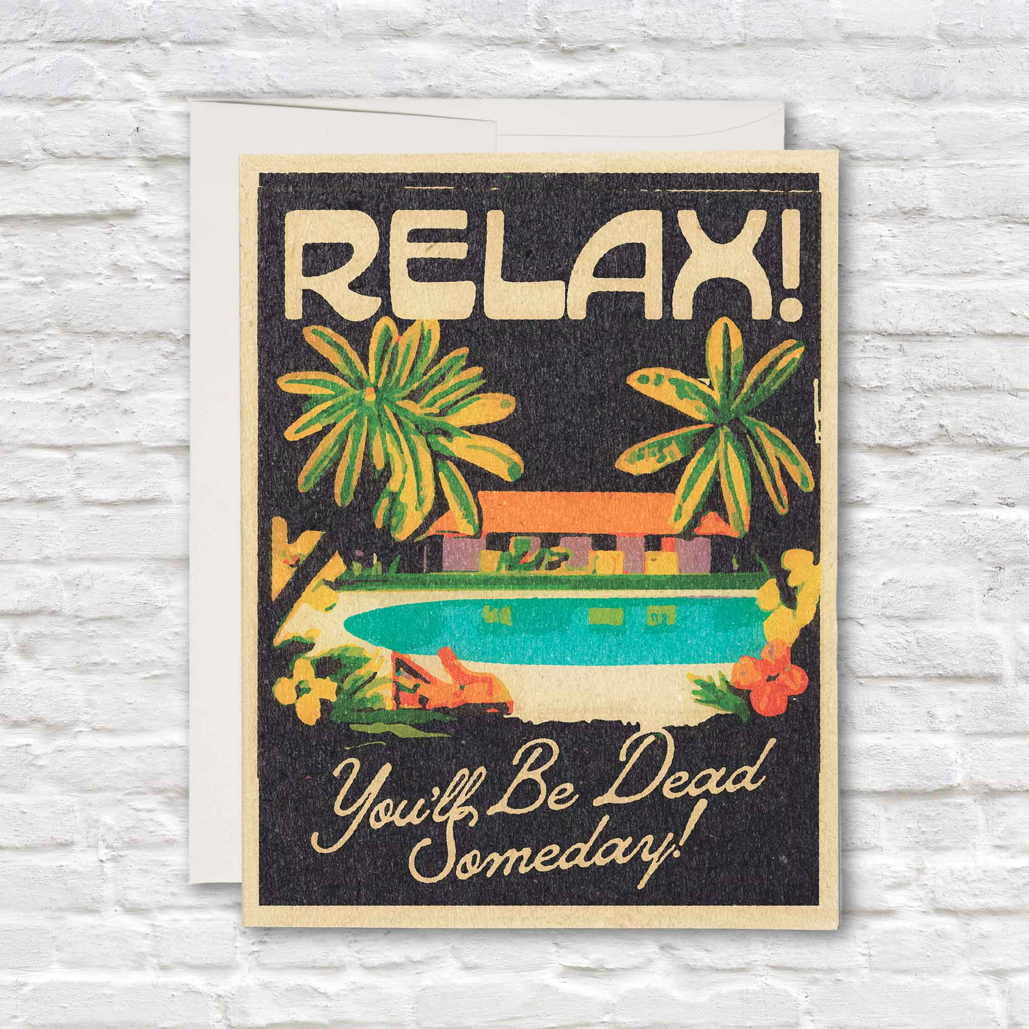 Red Cap Cards "Relax! You'll Be Dead Someday" greeting card illustrated by Daren Thomas Magee displayed with its matching envelope inside Gray Man Gallery in Pawleys Island, South Carolina. Humorous greeting card featuring a tropical pool, palm trees, and vintage-inspired typography.