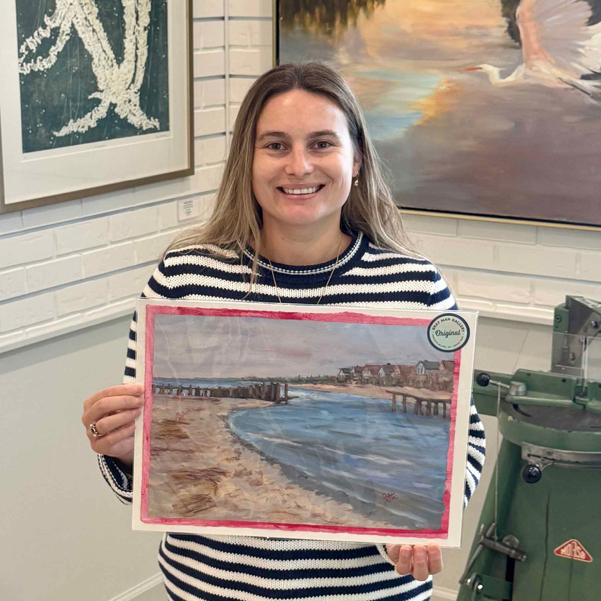 Gray Man Gallery team member holding Broken Groin South End #213, an original coastal oil on paper by local artist Retta Dotterer, showing its scale and distinctive pink painted border inside the Pawleys Island gallery.