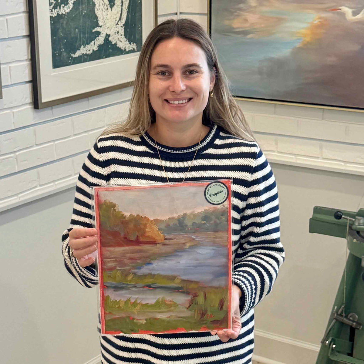 Gray Man Gallery team member holding Shem Creek #216, an original oil on paper by local artist Retta Dotterer, showing the scale and hand-painted coral edges inside the Pawleys Island gallery.